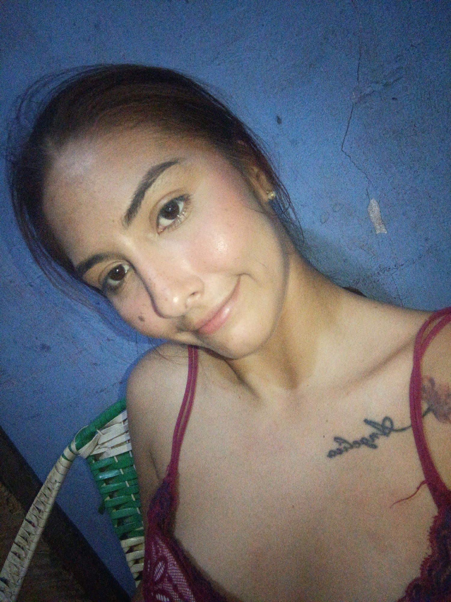 A woman with brown hair pulled back is looking at the camera with a slight smile. She has brown eyes and visible freckles on her face. She is wearing a red lace top with thin straps. She has a tattoo on her chest, and another tattoo on her upper right shoulder. She is sitting in a chair with green and white woven detailing. The background is a blue wall with some damage visible. There is no information to definitively ascertain this photo was taken in Juan Griego, Venezuela.