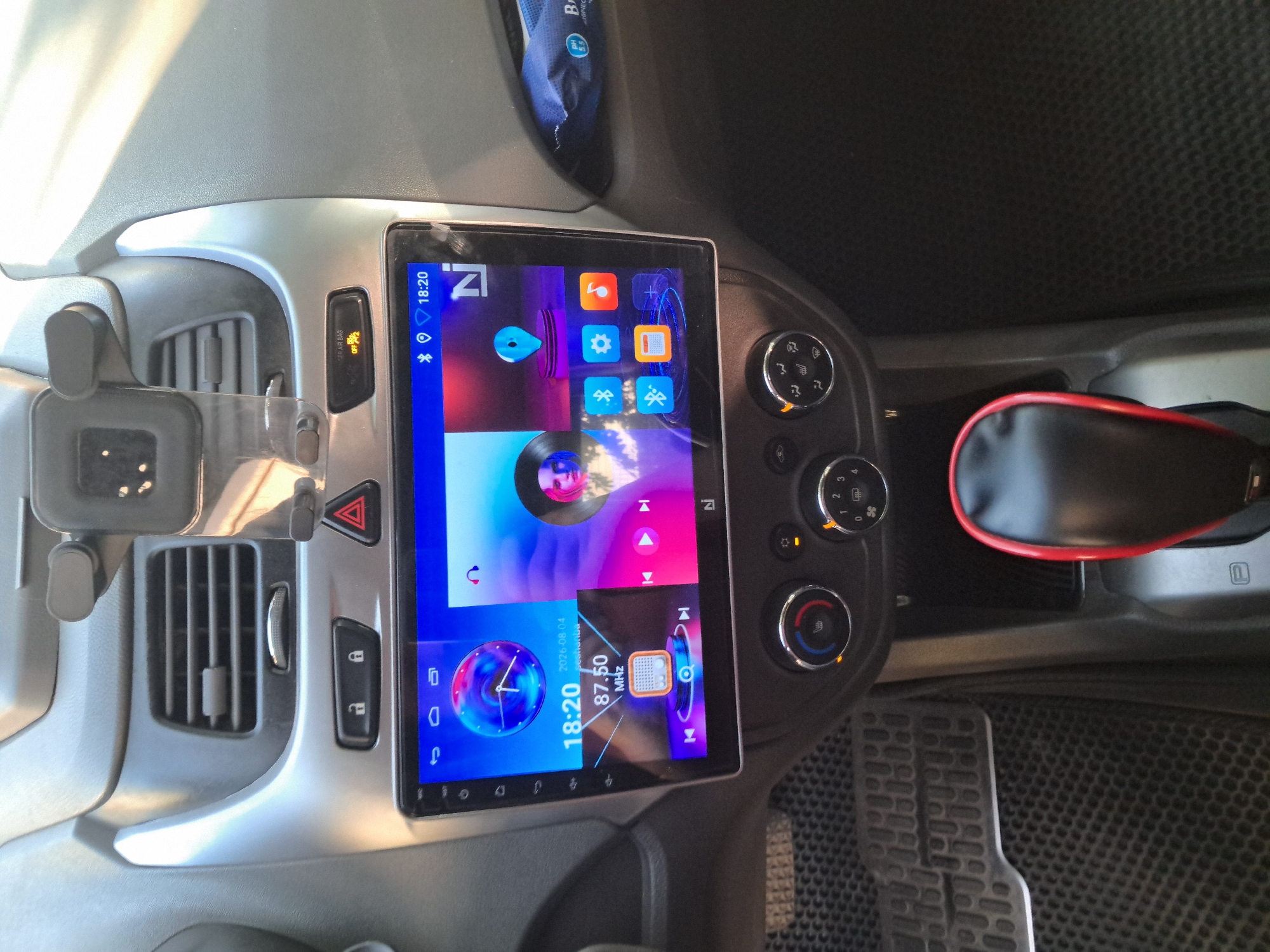 The image shows the interior of a car, specifically the dashboard and center console. A large touchscreen infotainment system is prominently displayed, showing various icons and information, including the time (18:20), date (2026.08.04), and what appears to be a radio frequency (87.50 MHz). The screen also displays a clock, a location pin icon, music controls, and a profile picture of a person. Below the screen are climate control knobs and buttons, as well as a gear shifter. The car's interior appears to be well-maintained. There are no people visible in the image. The setting is likely a private vehicle, not a public space. The provided context of "Bagish, Uzbekistan" is not visually verifiable from the image itself, as there are no landmarks or identifying features to place it within that specific city or country.