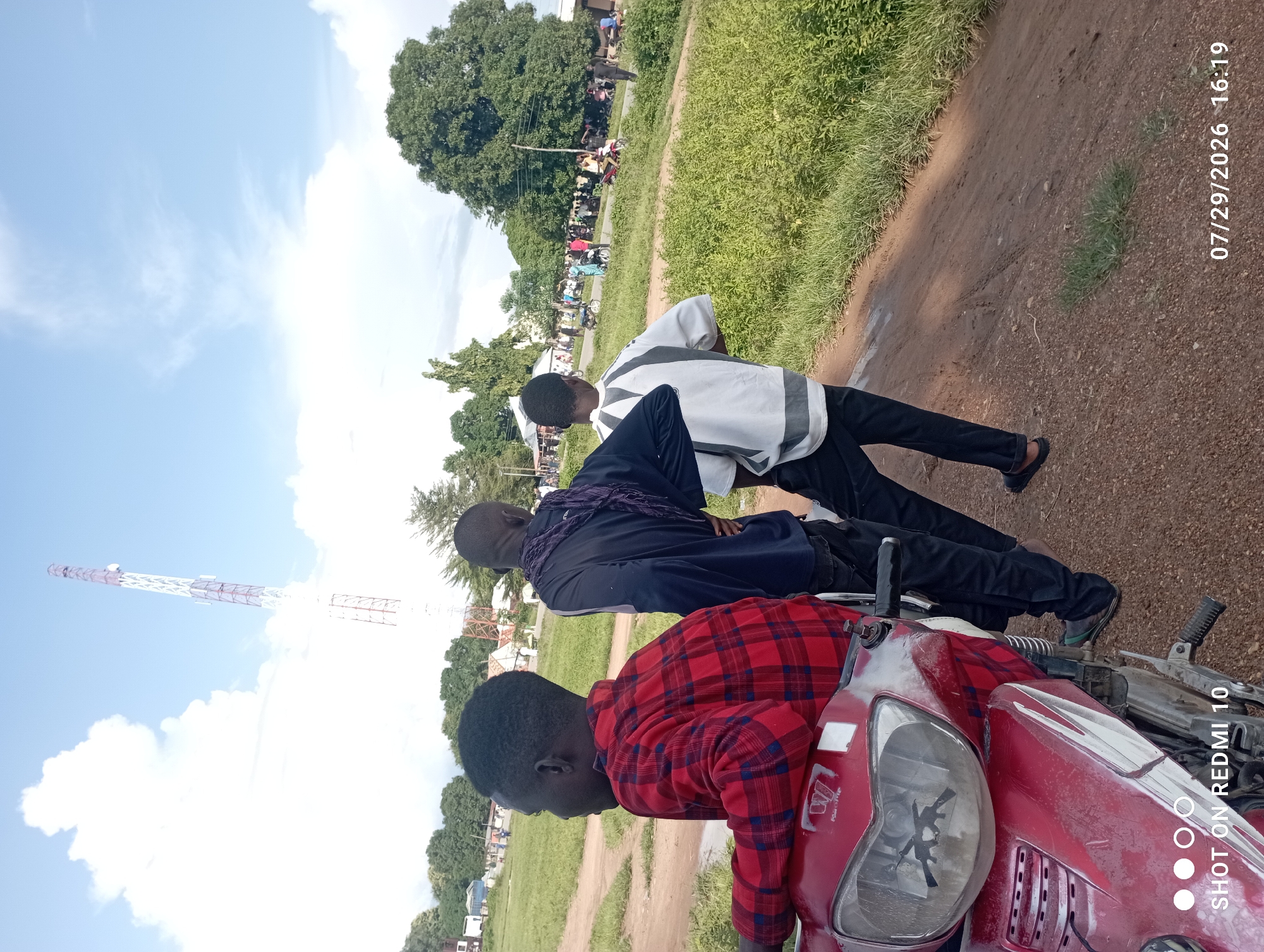 In the city of Serti, Nigeria, three young males are captured in a rear-view shot, positioned near a red motorcycle. The individual in the foreground wears a red and black plaid shirt and is seated on the motorcycle. To his left, another male is dressed in a dark blue long-sleeved shirt with a dark purple scarf draped around his neck. The third male stands to the right, attired in a white and grey long-sleeved shirt. The setting appears to be an outdoor area with a dirt road in the foreground, flanked by lush green grass and vegetation. In the background, a telecommunications tower is visible against a sky filled with white clouds and blue patches. Further in the distance, several people, motorcycles, and structures are present, suggesting a populated area or a gathering. The scene is illuminated by daylight, casting soft shadows. A timestamp overlay indicates the image was taken on 07/29/2026 at 16:19.