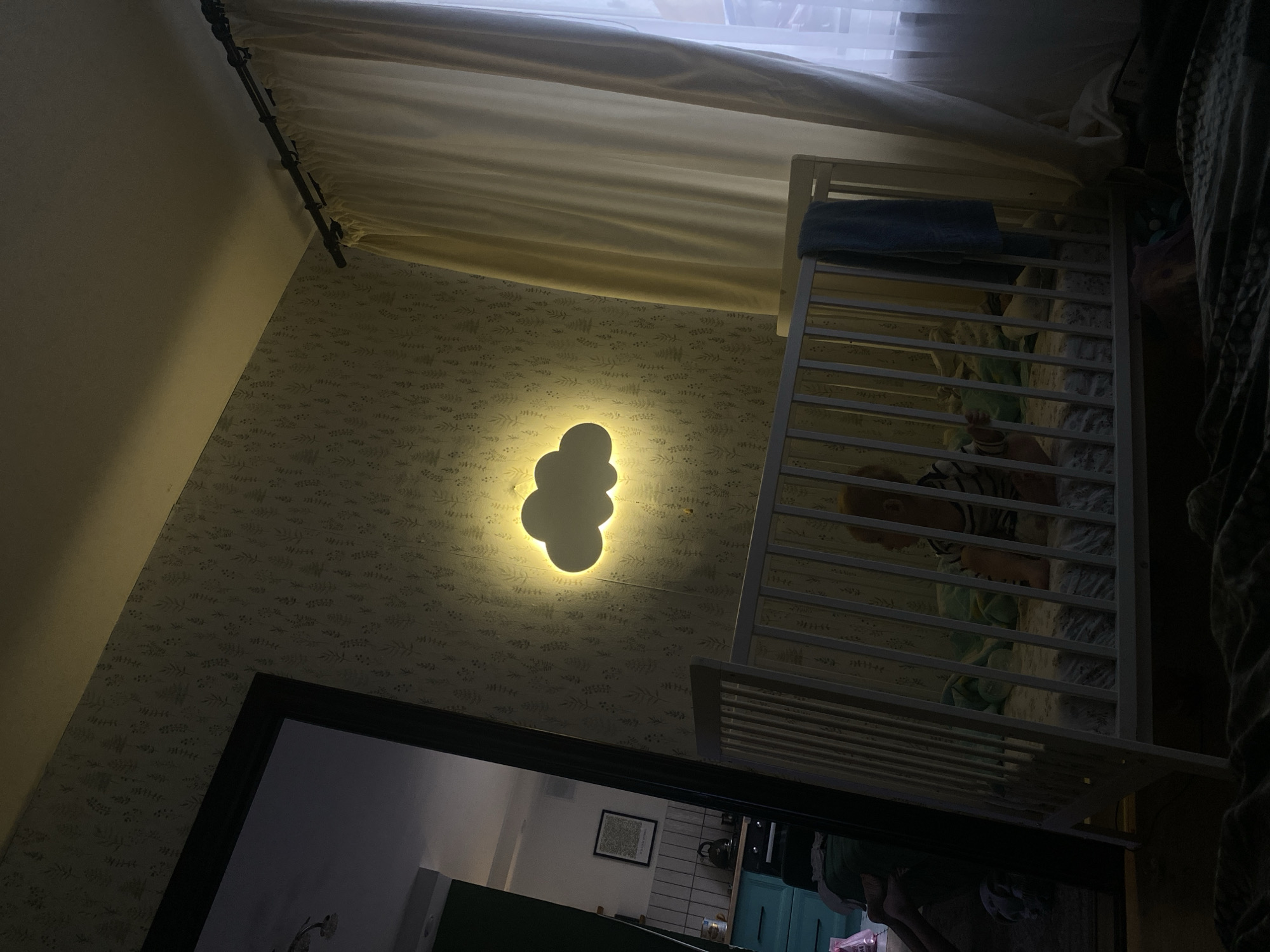 The image shows a room in Chaikovsky, Russia, featuring a child in a white crib with a blue towel draped over the top. The child is wearing a striped shirt and lying on a patterned blanket. A cloud-shaped light fixture is mounted on the wall covered in patterned wallpaper. There is a doorway leading to another room, which appears to be a kitchen, as evidenced by the teal cabinets and various kitchen appliances visible inside. Curtains hang from a rod near the ceiling.