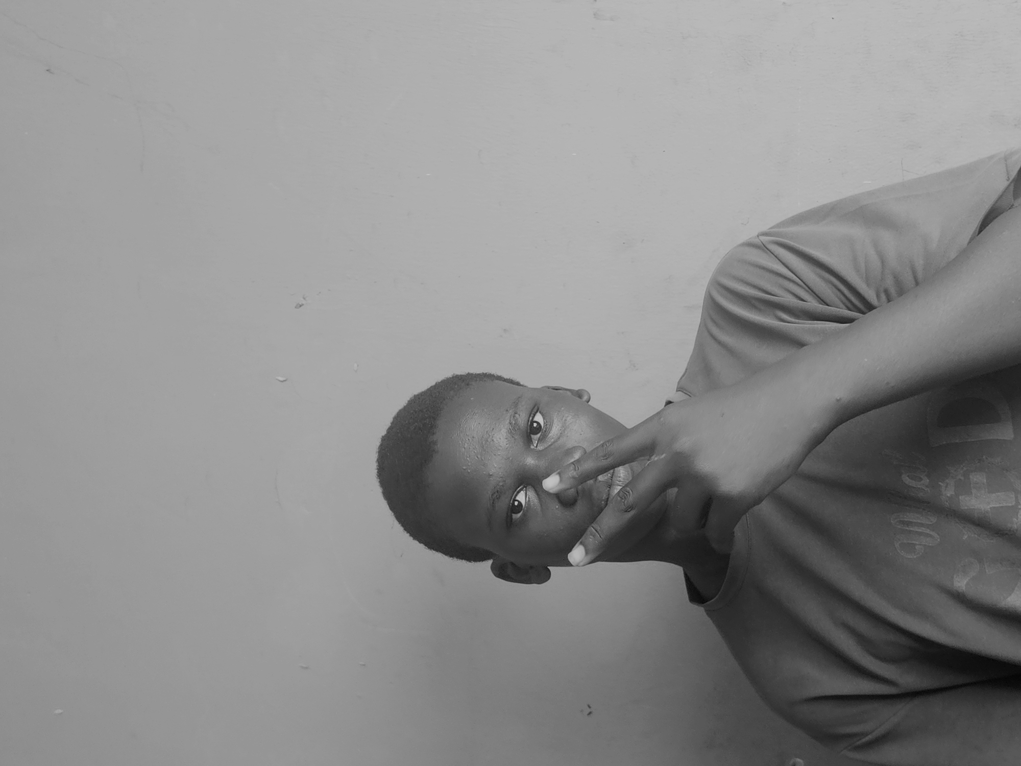 In this grayscale, medium shot, a young Black male is positioned centrally, facing forward with his head slightly tilted. He has short, dark hair and is looking directly at the camera. His right hand is raised, with the index and middle fingers extended in a V-shape, positioned in front of his face, near his nose and mouth. He is wearing a short-sleeved t-shirt with a visible graphic design on the chest. The background is a plain, textured wall, appearing to be an indoor setting with consistent lighting. The overall atmosphere is neutral and direct, highlighting the subject and his gesture.