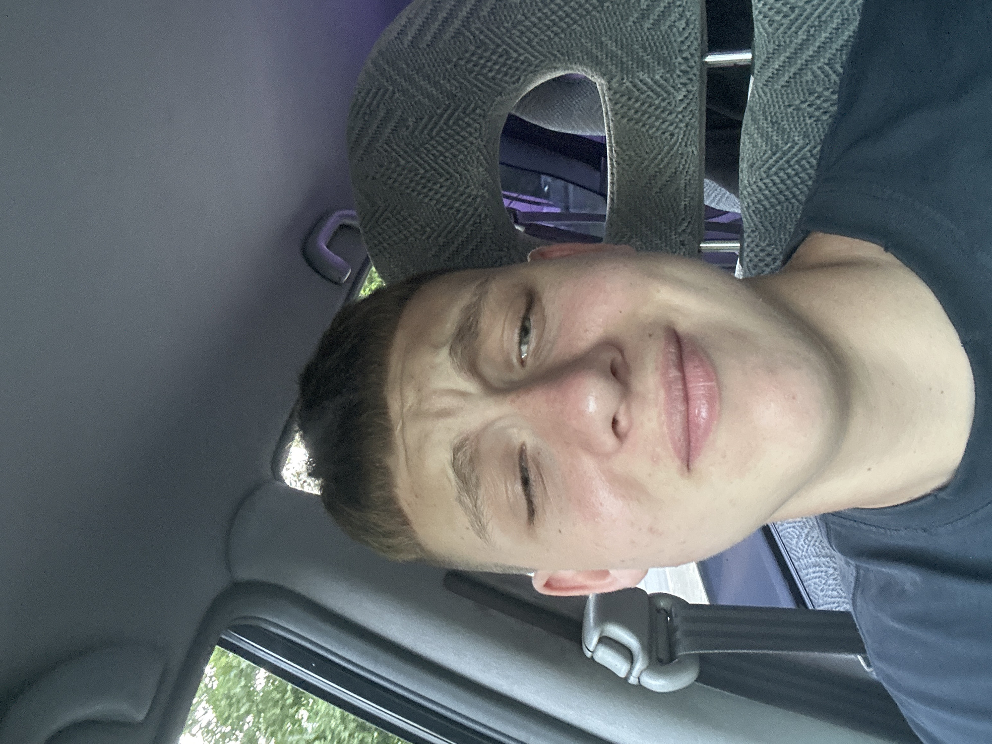 A young person with short, buzzed hair and fair skin is in the passenger seat of a car, looking into the camera. They are wearing a dark t-shirt. The interior of the car is visible, including the headrest of the seat behind them, which has a herringbone pattern. Sunlight filters in from the window on the left, illuminating trees and foliage outside. The overall lighting suggests it is daytime.