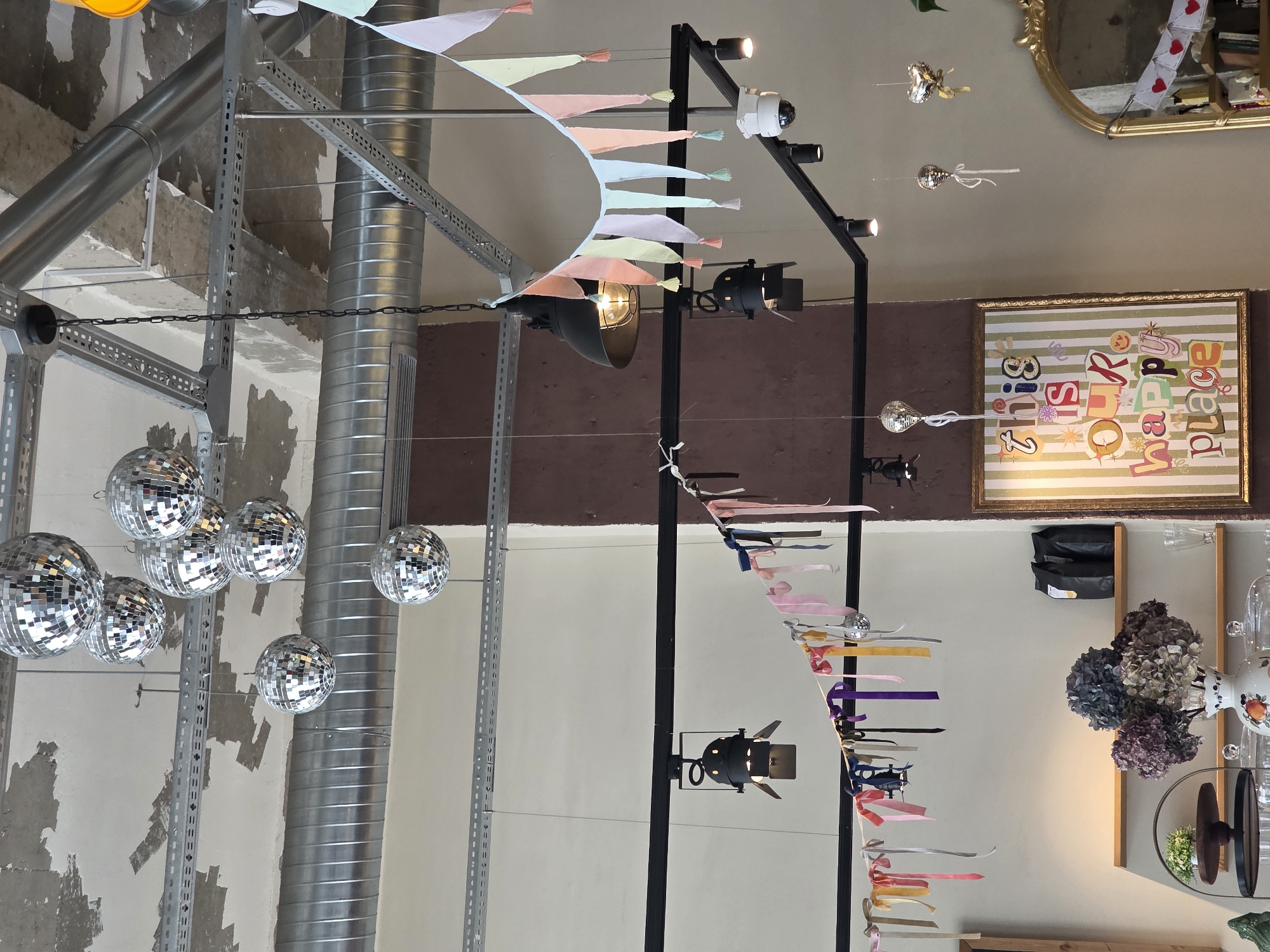 In the city of Khatay Raion, Azerbaijan, an interior scene is depicted, dominated by a rustic industrial aesthetic. The ceiling features exposed metal conduits and rough concrete, with several disco balls hanging at various heights. Below these, a black track lighting system is installed, from which additional spotlights and decorative elements are suspended.
A string of pastel-colored fabric pennants with tassels hangs across the scene, interspersed with ribbons of various colors tied to a black string. A surveillance camera is visible mounted on the track lighting system. In the lower portion of the image, a framed piece of art with the text "this is OUR happy place" is displayed, featuring colorful, collage-style lettering. Below the artwork and lighting, wooden shelves are present, holding black coffee bags, glassware, and a vase of dried hydrangeas. A decorative gold mirror is positioned to the right, with a string of red heart cutouts hanging in front of it. The overall ambiance suggests a contemporary establishment with a playful and celebratory theme.