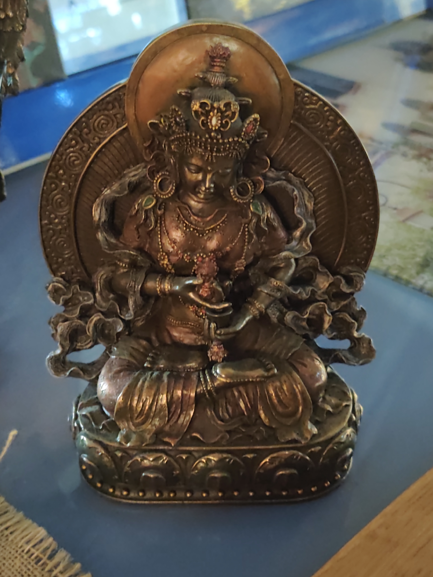 The image shows a close-up, high-angle shot of a bronze statue of a multi-armed deity, likely a Buddhist figure, seated on a decorative base. The statue is intricately detailed, with the deity adorned in elaborate jewelry, a crown, and flowing robes. One hand is outstretched, holding a small, reddish object, possibly a flower or rosary. The statue is positioned on a flat, blue surface, with a woven material and a wooden surface visible in the foreground and to the right, respectively. In the blurred background, a bright, illuminated object, possibly a screen or window with reflective properties, suggests an indoor setting. There is no indication of the city of Sochi, Russia, or any specific location within it. The scene is static and focuses solely on the statue.