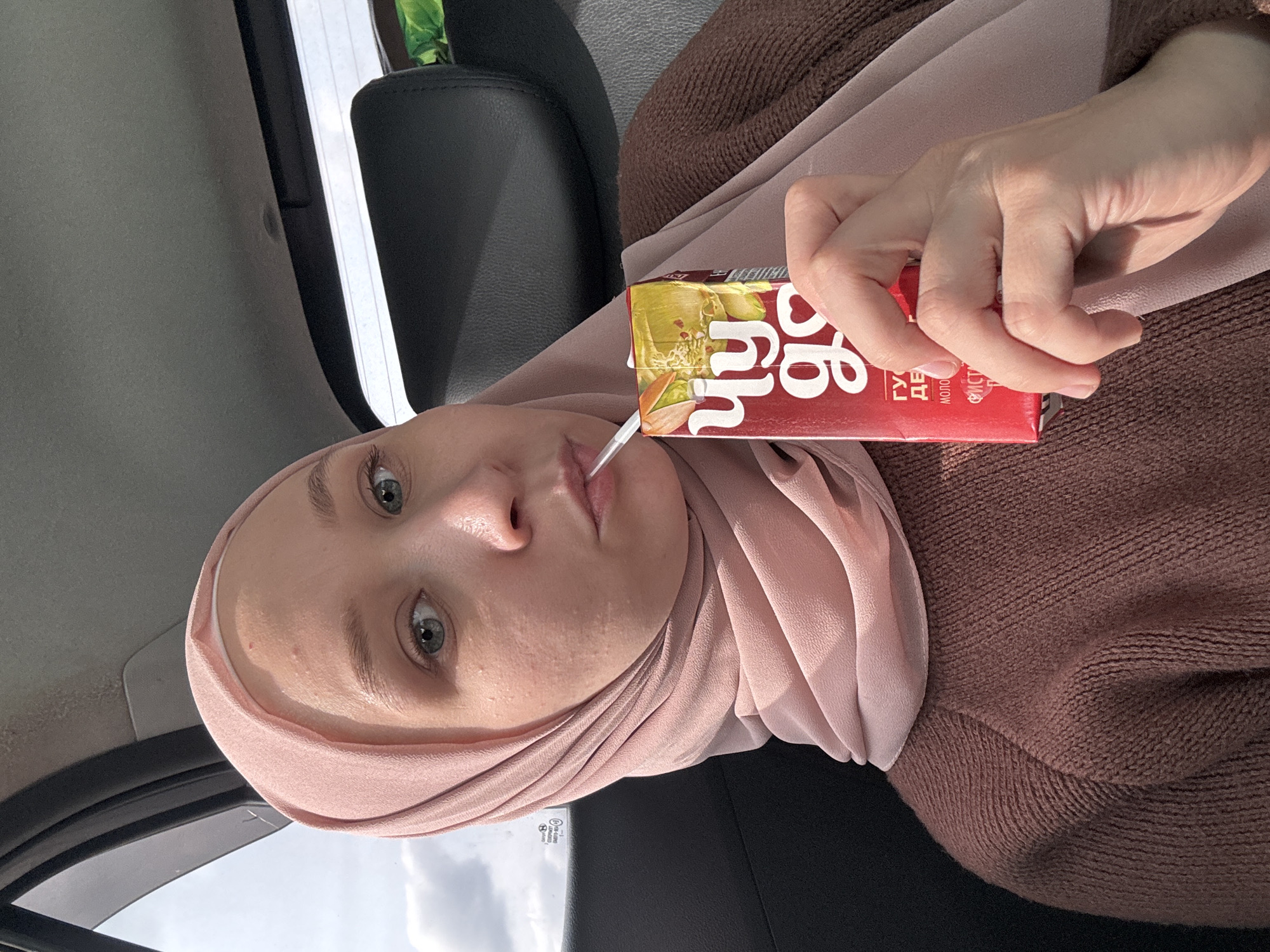 A woman wearing a pink hijab and a brown sweater is sitting in a car and drinking juice from a small carton using a straw. The juice carton is red, yellow, and white with writing in Cyrillic. The woman has fair skin and light eyes. The interior of the car is visible. The location may be Izhevsk, Russia.