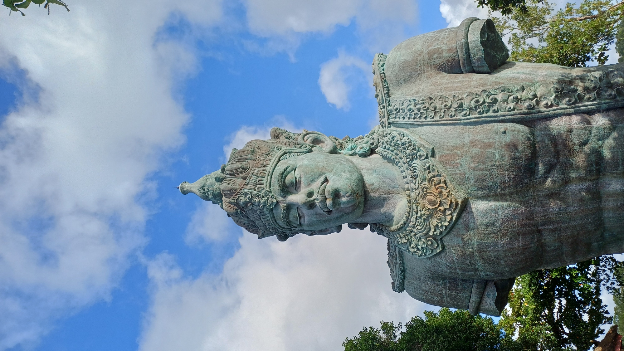 The image features a close-up view of a portion of the Garuda Wisnu Kencana statue, located in Kuta Selatan, Bali, Indonesia. The focus is on the head and upper torso of the statue, set against a backdrop of a partly cloudy sky. The statue appears to be constructed from a greenish-bronze-colored metal. The head is adorned with intricate carvings and details. The statue is viewed from below, looking upwards.