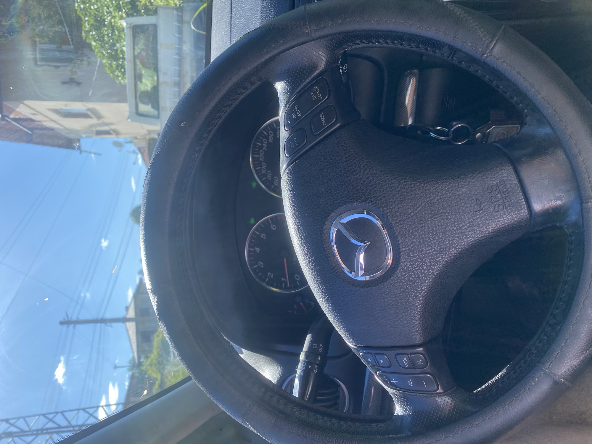 The image provides a first-person perspective from the driver's seat of a car. The steering wheel, partially covered by a black protective sleeve, is the central focus. The Mazda emblem is prominently displayed on the steering wheel. Various control buttons are visible on the steering wheel, including volume controls, mode selection, and cruise control buttons labeled "RES ACCEL," "CANCEL," and "SET COAST." The dashboard features a speedometer and tachometer, with the speedometer indicating a speed between 0 and 40 km/h. The exterior view through the windshield reveals a sunny day with a clear blue sky and scattered clouds. In the background, buildings and utility poles suggest a residential or semi-urban environment. The scene appears to be located in Sadovets, Bulgaria, as indicated by the presence of specific architectural styles and the overall atmosphere of a small town or village. A white pickup truck is visible parked in the distance. The overall impression is that of a paused moment, perhaps during a drive or a stop in Sadovets.