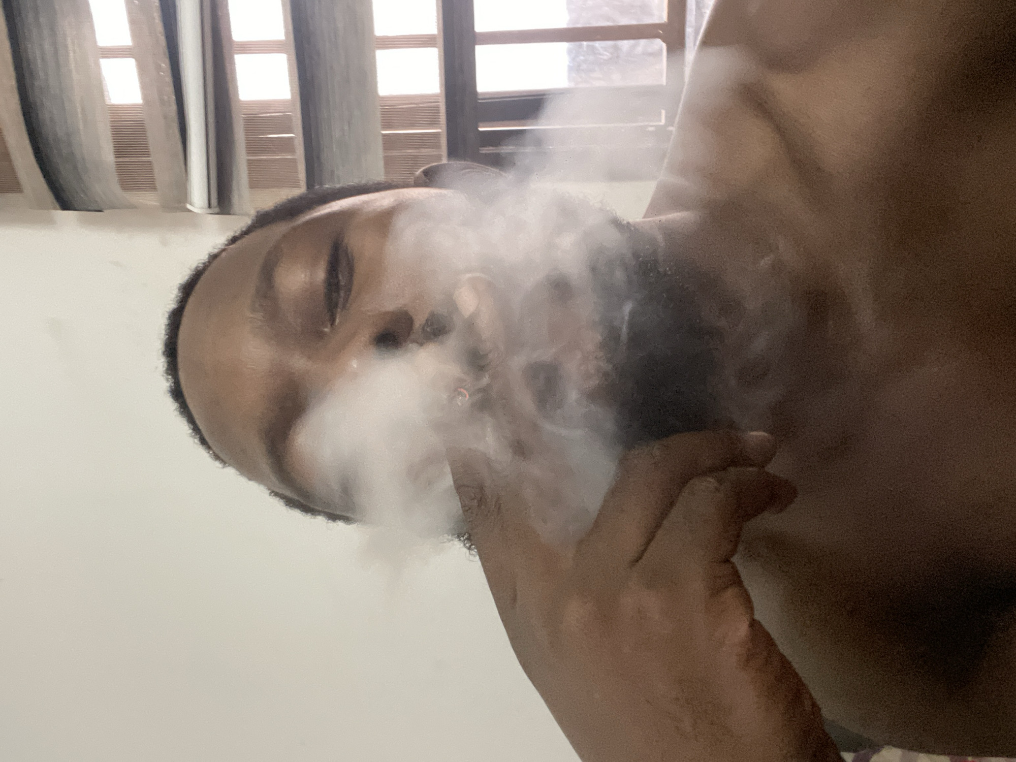 The image displays a close-up, rotated portrait of a shirtless Black man in an indoor setting, likely in Abuja, Nigeria, during the daytime. The man, who has a beard, is positioned horizontally across the frame, with his head on the left and his body extending to the right. A prominent cloud of white vapour or smoke emanates from and surrounds his mouth and nose, suggesting he is exhaling from vaping or smoking. His eyes appear closed or heavily lidded, conveying a sense of relaxation or focus on the act.
In the background, light-colored curtains cover a window, through which natural light filters, indicating it is daytime. The window itself has horizontal blinds or louvres. The man's skin is dark, and his upper body is visible, showing his shoulder and chest area. The overall lighting is soft, primarily coming from the window. The dominant colors are the man's skin tones, the white vapour, and the light grays and whites of the curtains and the surface beneath him. No text is visible in the image.