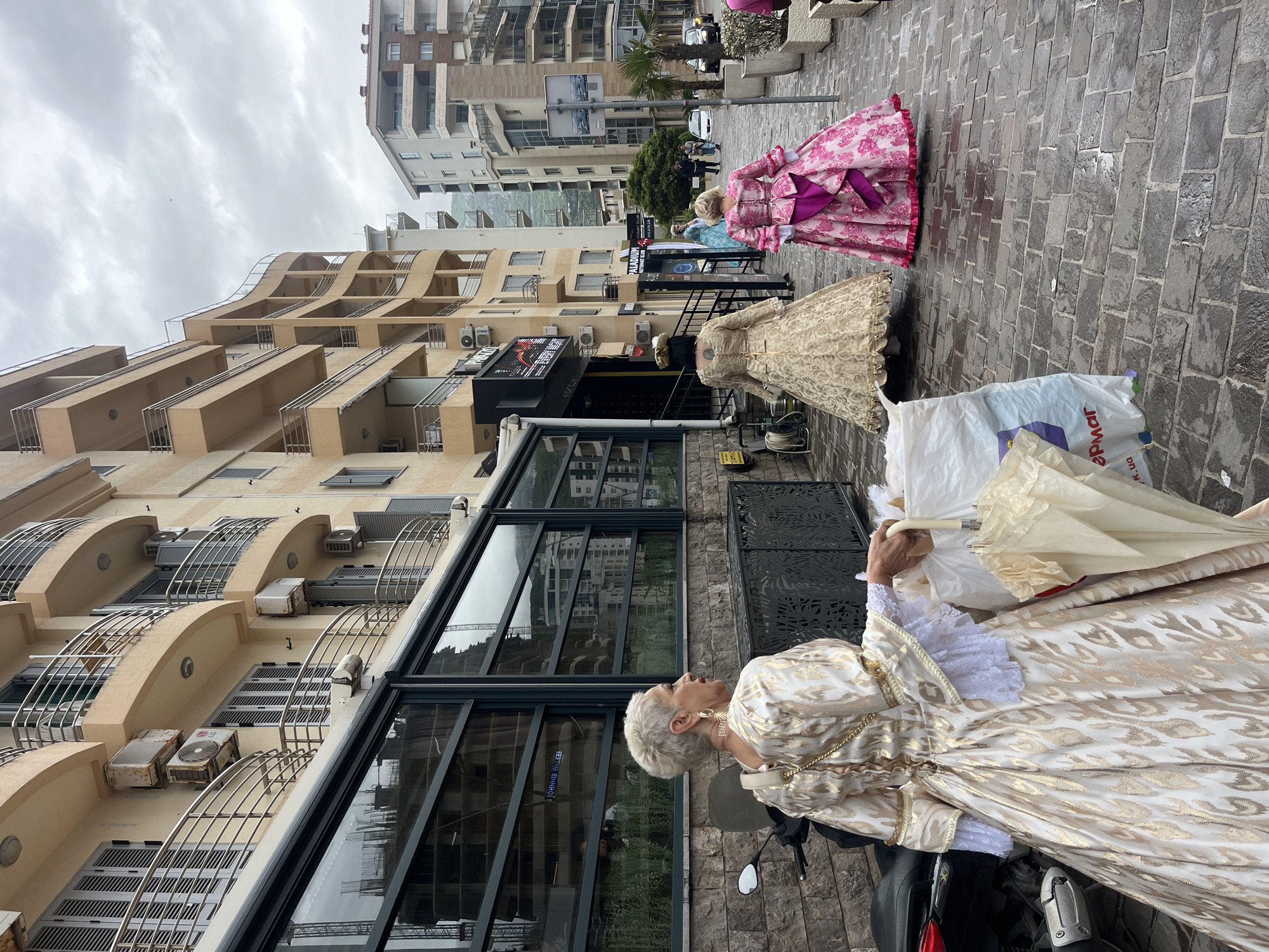 The image depicts a scene in Budva, Montenegro, featuring three individuals dressed in ornate, historical-style gowns. The foreground shows a person in a gold and white dress, holding a closed white parasol. They are seated on a black motor scooter, which is parked in front of a modern building with large, dark-framed windows. The middle ground includes two mannequins in elaborate dresses; one is gold, and the other is pink. These mannequins are positioned on the brick-paved sidewalk along with a large white shopping bag. The background displays modern, multi-story buildings with balconies and air conditioning units, typical of an urban area. The sky is overcast, and the street appears wet, suggesting recent rain. In the distance, beyond the mannequins, there are hints of traffic and other pedestrians. The scene suggests a possible theatrical event or a costume-related activity.