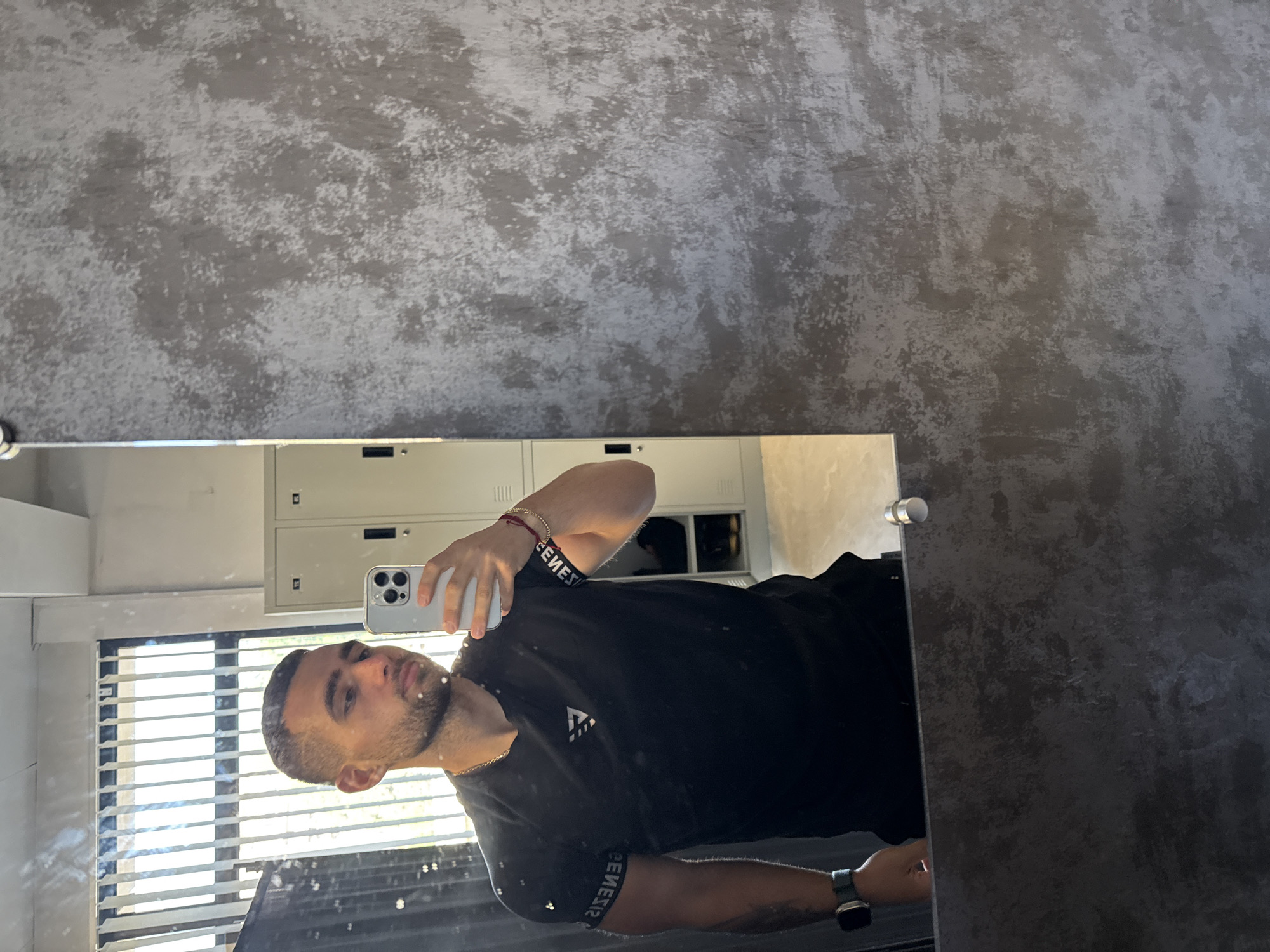 A young man with short, dark hair and a beard is taking a mirror selfie in what appears to be a locker room. He is wearing a black t-shirt with a white logo on the chest and a white band with black lettering on his left bicep. He is also wearing a silver chain necklace and a watch on his left wrist. The man is holding a silver smartphone in his right hand, angled to capture his reflection. In the background, there are several light-colored lockers with black handles. A window with vertical blinds is visible to the left of the lockers, allowing natural light to enter the room. The walls have a textured, grey, concrete-like finish.