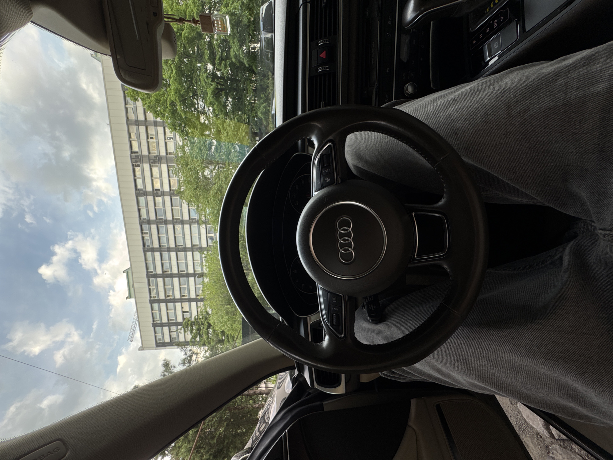 The image shows the interior of an Audi car, likely taken from the driver's perspective. The steering wheel, bearing the Audi logo, is prominent. A person wearing gray denim pants is sitting in the driver's seat. Outside the car window, a modern building with numerous windows is visible, suggesting an urban environment. The location appears to be Tbilisi, Georgia. Trees are visible interspersed with the building. The sky is partly cloudy.