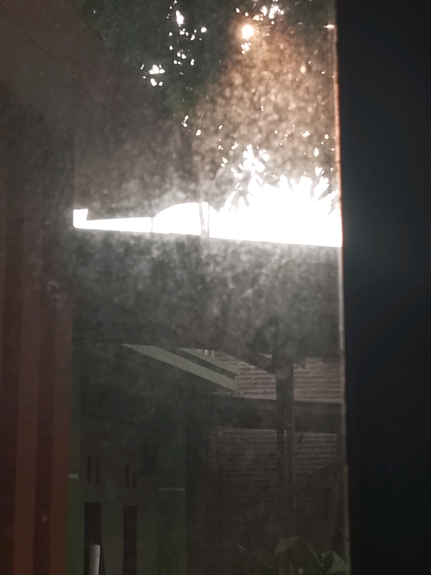 A reflection on a dusty window shows sunlight filtering through leaves, creating bright spots. Below the sunlight, the reflection reveals a glimpse of outdoor structures, including a brick wall and wooden beams, suggesting an urban or semi-urban environment. The foreground is dominated by the textured surface of the window, with its smudges and particles obscuring a clearer view of the reflected scene. The overall impression is one of looking out from a sheltered interior, possibly a home, onto an obscured outdoor space. The location in Ujungrusi, Indonesia, implies a tropical or subtropical setting, consistent with the foliage visible in the reflection.