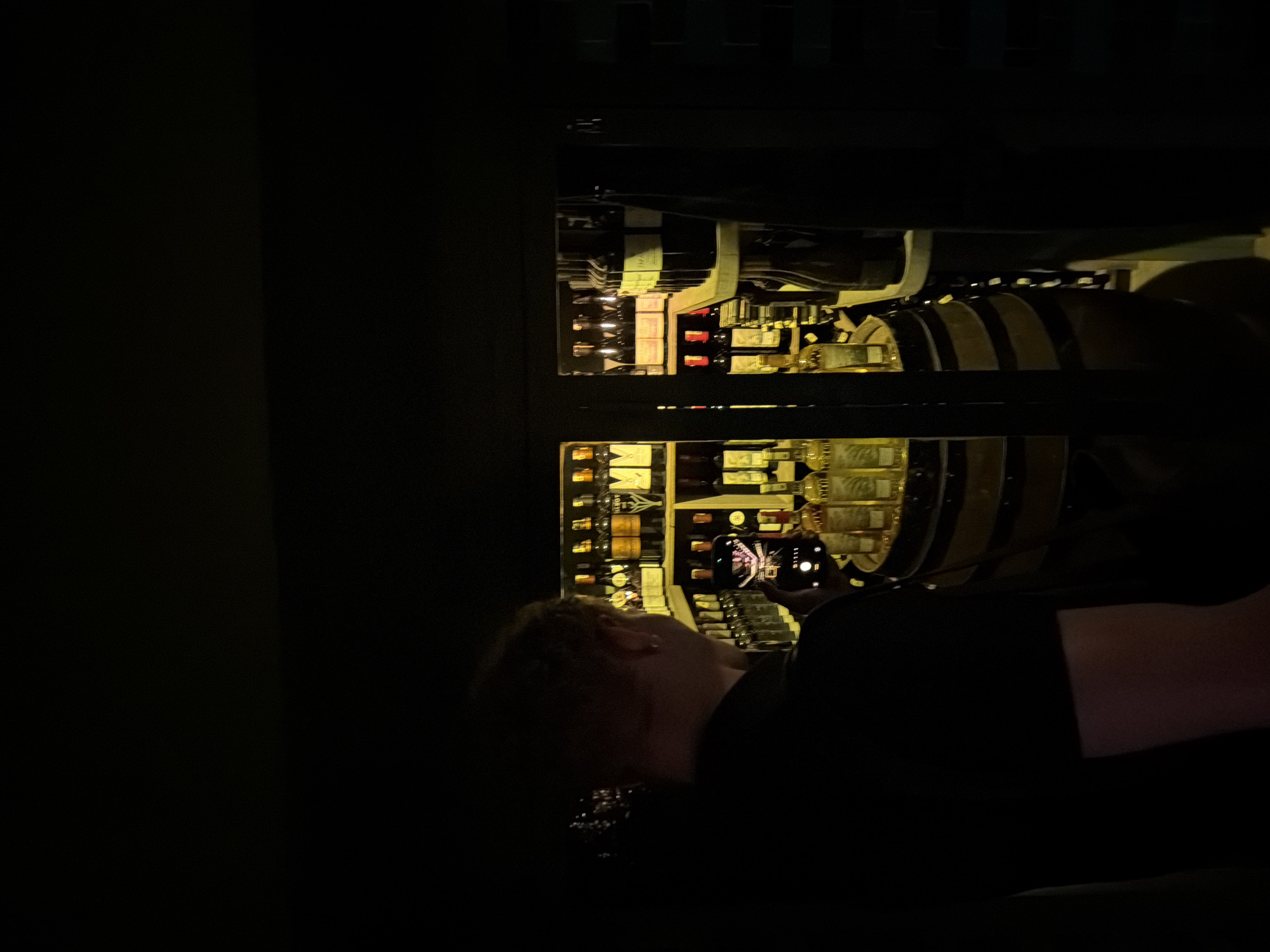 The image captures a dimly lit scene, likely inside a wine cellar or a specialized shop in Marrakesh, Morocco. A person, seen from the back and to the left, is holding a smartphone, appearing to take a photograph or video. They are wearing a dark t-shirt. In front of the person is a large wooden barrel, upon which several bottles are displayed. Behind the barrel, shelves are packed with numerous bottles of wine, illuminated by warm, ambient lighting that casts yellow hues. The shelves are organized and well-stocked, suggesting a curated collection. The overall atmosphere is intimate and perhaps slightly mysterious due to the low light conditions. The architecture and style of the display, with the barrel and numerous wine bottles, are consistent with a sophisticated establishment that might be found in a vibrant city like Marrakesh, known for its diverse offerings.