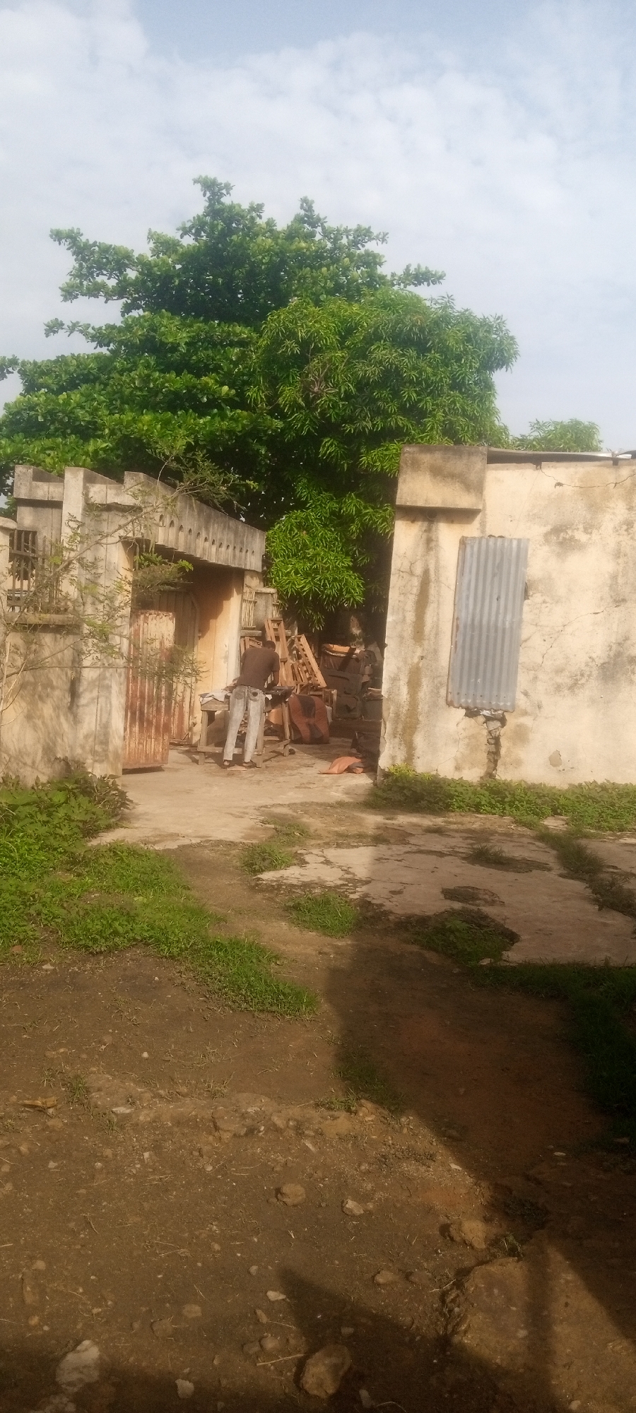 The image depicts an outdoor scene in Sabon Tasha, Nigeria. A man is working in the yard of an old, weathered structure that appears to be a workshop or residence. The building is constructed of concrete blocks. A rusted gate sits next to the man. The ground is a mix of dirt and patches of grass. The sky is overcast. A large tree is overhead.