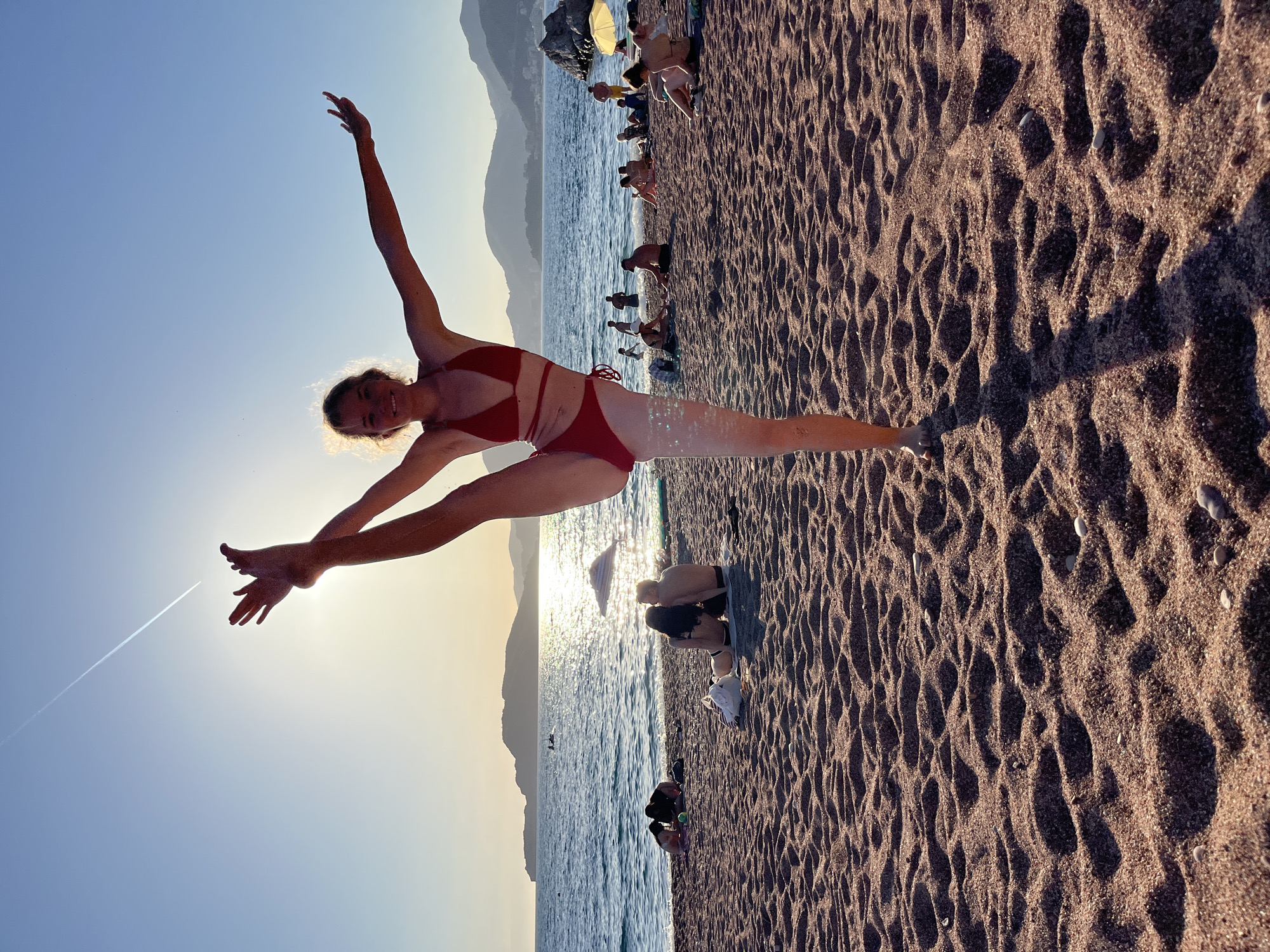 The image depicts a vibrant beach scene at sunset in Rađenovići, Montenegro, characterized by warm light and numerous individuals. In the foreground, a young female with blonde, curly hair, wearing a red two-piece bikini, strikes a dynamic yoga or gymnastic pose. She stands on her right leg, which is subtly adorned with shimmering specks, while her left leg is extended vertically upwards, with her left hand grasping her left foot. Her right arm is outstretched to the side for balance, and she smiles directly at the viewer. The setting sun, positioned behind her head and upper torso, creates a strong backlight, forming a bright halo around her hair and highlighting the contours of her body, casting a distinct shadow on the sandy beach in front of her.
The beach itself is composed of reddish-brown sand, marked by numerous footprints and scattered small pebbles, indicative of recent activity. In the midground, the beach is populated by several people engaged in various leisure activities. A couple sits closely together on a white towel near the water's edge, facing the sea. Other individuals are either relaxing on the sand, wading, or standing in the shallow, calm blue water of the Adriatic Sea. Visible objects include a striped beach umbrella, a yellow beach umbrella further right, and what appears to be a surfboard lying near the water.
The background features the calm sea, reflecting the golden light of the setting sun in a shimmering path across its surface. Distant mountains or hills are silhouetted against the horizon, with the landmass extending to the left. The sky is a clear gradient of blue, transitioning to lighter, warmer tones near the horizon. A prominent, white aircraft contrail extends across the upper left portion of the sky.