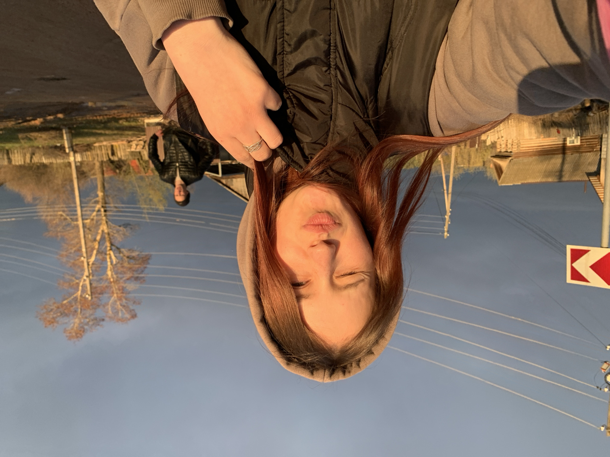 An outdoor, slightly off-kilter selfie captures a young woman in the foreground and a rural setting in the background. The woman, with reddish-brown hair, is wearing a grey hooded sweatshirt and a black puffer vest. She looks towards the camera with her eyes closed and lips pursed, an expression indicative of a playful moment. A ring is visible on her left hand.
Behind her, to the left, a man wearing a dark jacket and a beanie walks along what appears to be a dirt or gravel road, partially obscured and slightly out of focus. The scene unfolds under a clear blue sky during the golden hour, with warm, low sunlight casting a glow on the subjects and environment. Bare-branched trees, suggesting late autumn or early spring, are scattered throughout the background, along with wooden fences and structures typical of a village or countryside setting in Foky, Russia. Overhead, multiple power lines crisscross the frame. On the far right, a red and white chevron road sign, indicating a sharp turn or hazard, is partially visible.