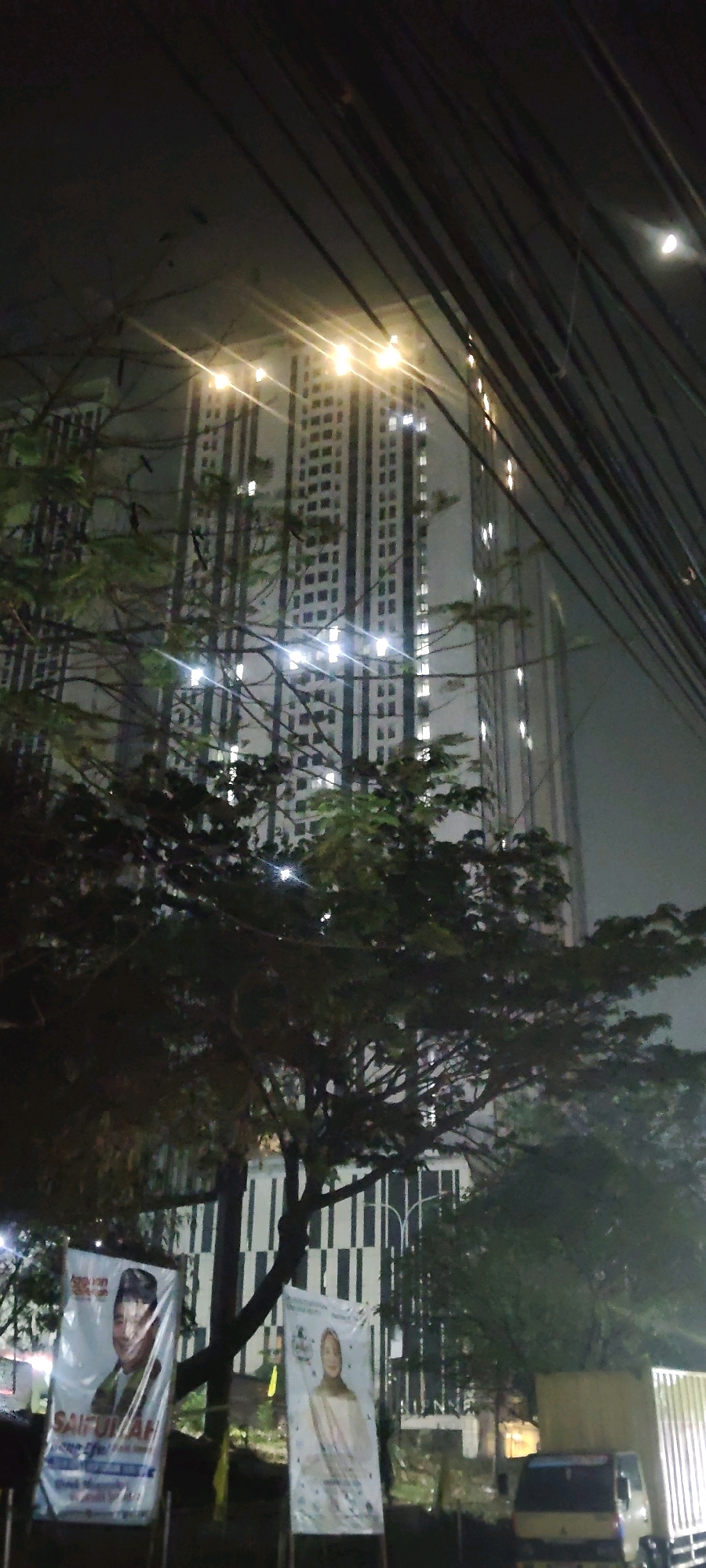 In Sukaresmi, Indonesia, at night, a tall apartment building with many lit windows is visible in the background, partially obscured by tree branches and overhead power lines. In the foreground, two banners are displayed. The banner on the left features a portrait of a man wearing a cap and a green shirt, with text below that includes "SAIFULLAH." The banner on the right shows a portrait of a woman wearing a headscarf and a cream-colored outfit. A yellow truck is parked in the right portion of the frame. The overall scene is dark and atmospheric, with the lights from the building and streetlights illuminating the surroundings.