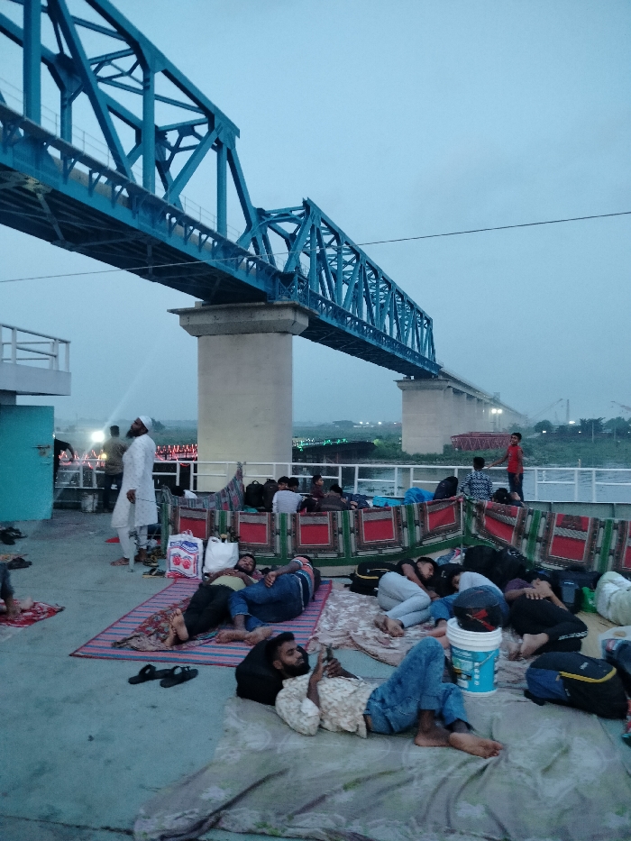 The image shows a group of people relaxing on the roof of a ferry under a large blue steel bridge. The ferry is docked and the riverbank is visible beyond the bridge. The location is Zazira, Bangladesh, at coordinates (latitude: 23.649, longitude: 90.464). The people are mostly lying down or sitting on colorful blankets. Some are talking, others are sleeping, and one man is looking at his phone. The bridge is a prominent feature in the image, with its blue steel structure standing out against the pale sky. The environment is a mix of urban and natural, with the bridge and ferry representing urban elements and the riverbank representing the natural environment. The image conveys a sense of tranquility and everyday life on the river.