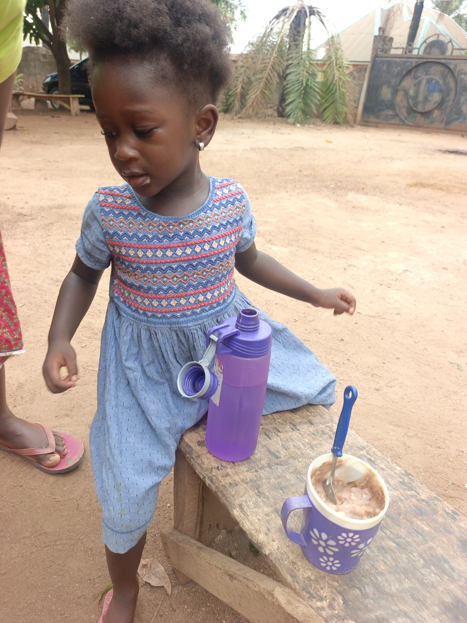 A young child with dark skin and short, curly hair stands near a wooden bench in what appears to be an outdoor setting. The child is wearing a blue dress with a patterned bodice and is looking down. There is a purple water bottle with an attached lid, and a purple mug with white flowers containing a food item and a blue spoon, both placed on the bench. A person wearing red patterned cloth and pink flip-flops stands in the background, partially cropped from the image. The ground is reddish-brown dirt. In the background, there is vegetation, a car, a wooden structure, and a metal gate. The location is indicated as Akwanga, Nigeria.