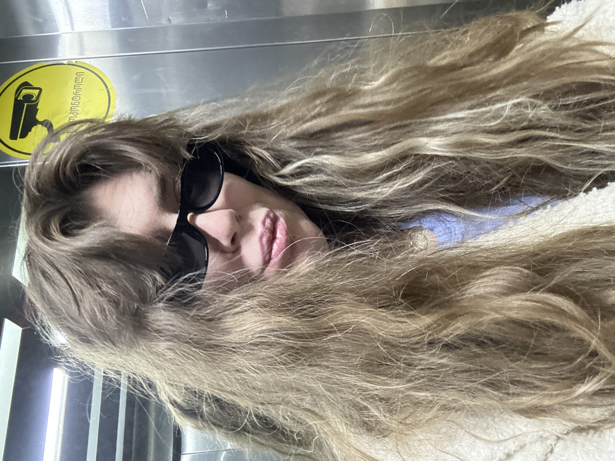 The image features a person, likely female, with long, wavy, light brown and blonde hair cascading across the frame. She is wearing black, rectangular sunglasses that cover her eyes, and her lips are slightly parted. Her attire includes a light-colored, possibly fluffy, outer garment over a blue top. The background consists of metallic, possibly stainless steel, panels and some vertical light sources on the left side, suggesting an indoor, modern, or industrial setting.
In the upper left corner, a yellow circular sticker is clearly visible. It displays a black icon of a security camera and Georgian text. The text reads "ვიდეომეთვალყურეობა" (videometvalyureoba), which translates to "video surveillance." This sign indicates that the area is under camera monitoring. The overall scene suggests the person is posing for a photograph in a public or semi-public indoor space, consistent with the provided location context of Tbilisi, Georgia. There are no specific cues regarding the time of day or weather.