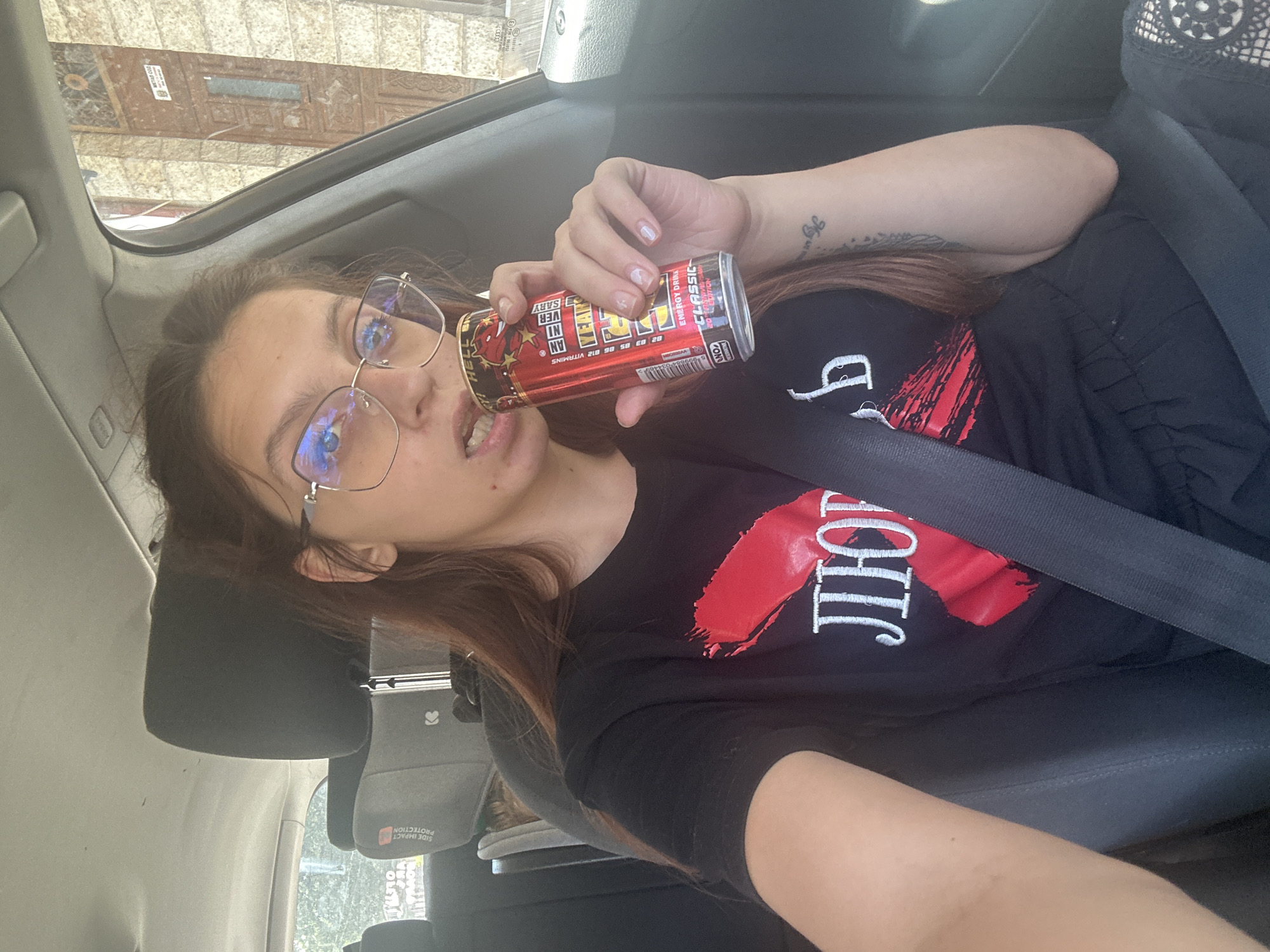 A woman with long brown hair, wearing glasses and a black t-shirt with a red design and white lettering, is sitting in the driver's seat of a car and drinking from a red energy drink can. The car appears to be parked on a street in the city of Tryavna, Bulgaria, with a building visible through the passenger window. The woman is wearing a seatbelt, and the car's interior is visible, including the headrest and part of the dashboard.
