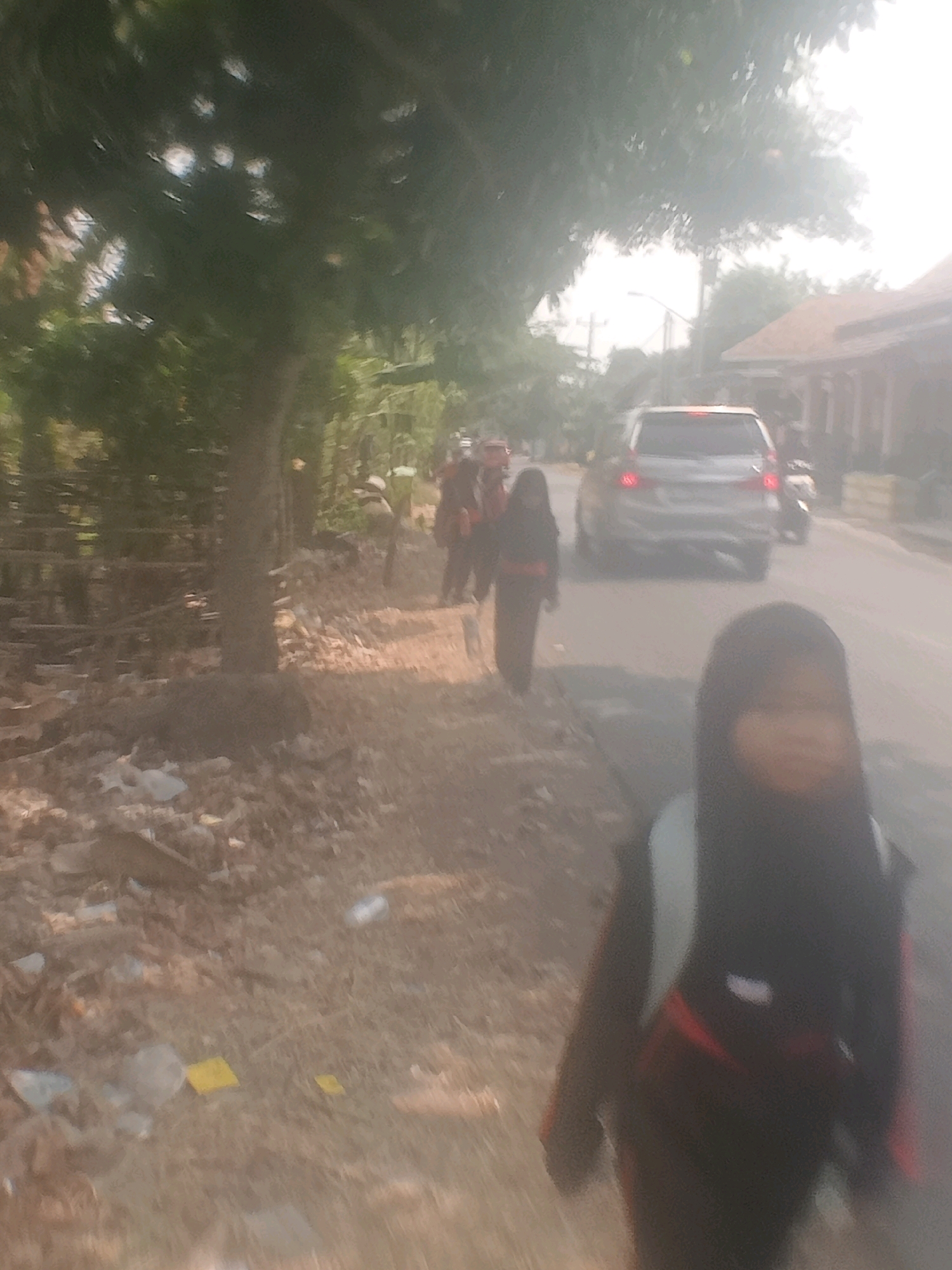 The image depicts a street scene in Lebakgowah, Indonesia. In the foreground, a young person wearing a dark headscarf and a backpack is visible, facing away from the camera. To the left, a dirt path is lined with trees and scattered debris, including plastic bottles and papers. A person in dark clothing is seen walking on the path in the middle ground. Further down the street, a silver SUV with its taillights illuminated is driving away from the camera. Several other individuals, appearing to be children, are walking along the side of the road. The background shows houses with tiled roofs and a bright, hazy sky, suggesting a sunny day. The overall atmosphere is casual, with people moving along a street that appears to be in a suburban or rural part of the city.