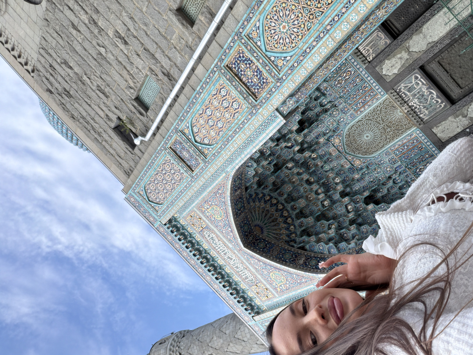 In Saint Petersburg, Russia, a young woman with long brown hair is taking a selfie in front of the vibrant, intricately tiled facade of the Saint Petersburg Mosque. The mosque, a prominent landmark, features a large arched entrance adorned with geometric patterns and Arabic calligraphy in shades of blue, teal, and gold. Above the entrance, the building's stone walls are visible, leading up to a patterned dome. A minaret is also partially visible in the background. The woman is wearing a white textured top with ruffled details and a gold ring on her right hand, which she is touching to her cheek. The sky above is a pale blue with scattered white clouds. The image is framed with a low angle, emphasizing the architectural grandeur of the mosque.