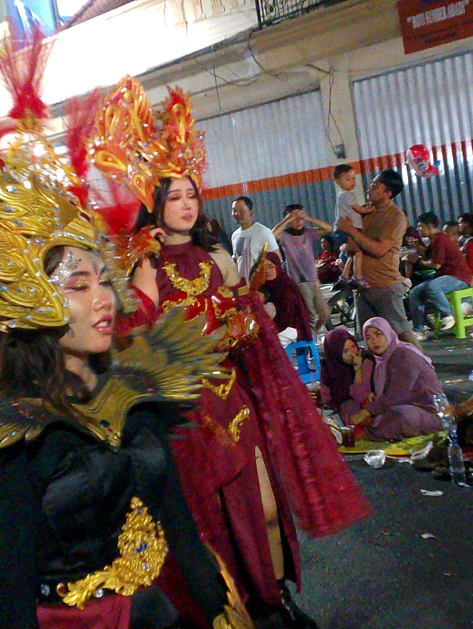 The image depicts a street scene in Probolinggo, Indonesia, likely during a cultural event or parade. In the foreground, two women are prominently featured, adorned in elaborate, colorful costumes and headdresses. The woman on the left wears a gold and black outfit with a feathered shoulder piece and a large gold headdress. The woman on the right is dressed in a maroon costume with a striking red and gold fiery headdress. Both women have distinct makeup, suggesting they are performers or participants in a pageant.
Behind them, a crowd of onlookers is visible. Several individuals are dressed in casual attire, while others are seated on the ground or on plastic stools. Among the spectators, a man is holding a young child, and there are other people observing the event. The background reveals a street environment with buildings, a corrugated metal wall, and some debris on the ground, including plastic bottles and wrappers. The lighting suggests it is nighttime or dusk, with artificial lights illuminating the scene. A red balloon is visible in the upper right portion of the image. The overall atmosphere appears to be festive and communal.