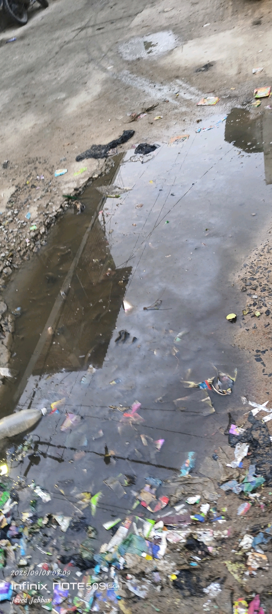 The image depicts a flooded street in the city of Bahera, India, littered with an assortment of trash. The waterlogged area shows a muddy ground with a significant accumulation of debris, including plastic wrappers, paper, and other miscellaneous waste. The water reflects the overcast sky, creating a dull and somewhat gloomy atmosphere. A portion of a motorcycle's wheel is visible in the upper left corner of the image, suggesting it is parked nearby. The presence of a date and time stamp, "2026/09/08 08:09," along with "Infinix NOTE 50S 5G" and "Javed Jabbar," indicates that the photograph was taken with a mobile device and possibly captured by Javed Jabbar. The scene highlights a common urban environmental issue of poor waste management and sanitation in the area.