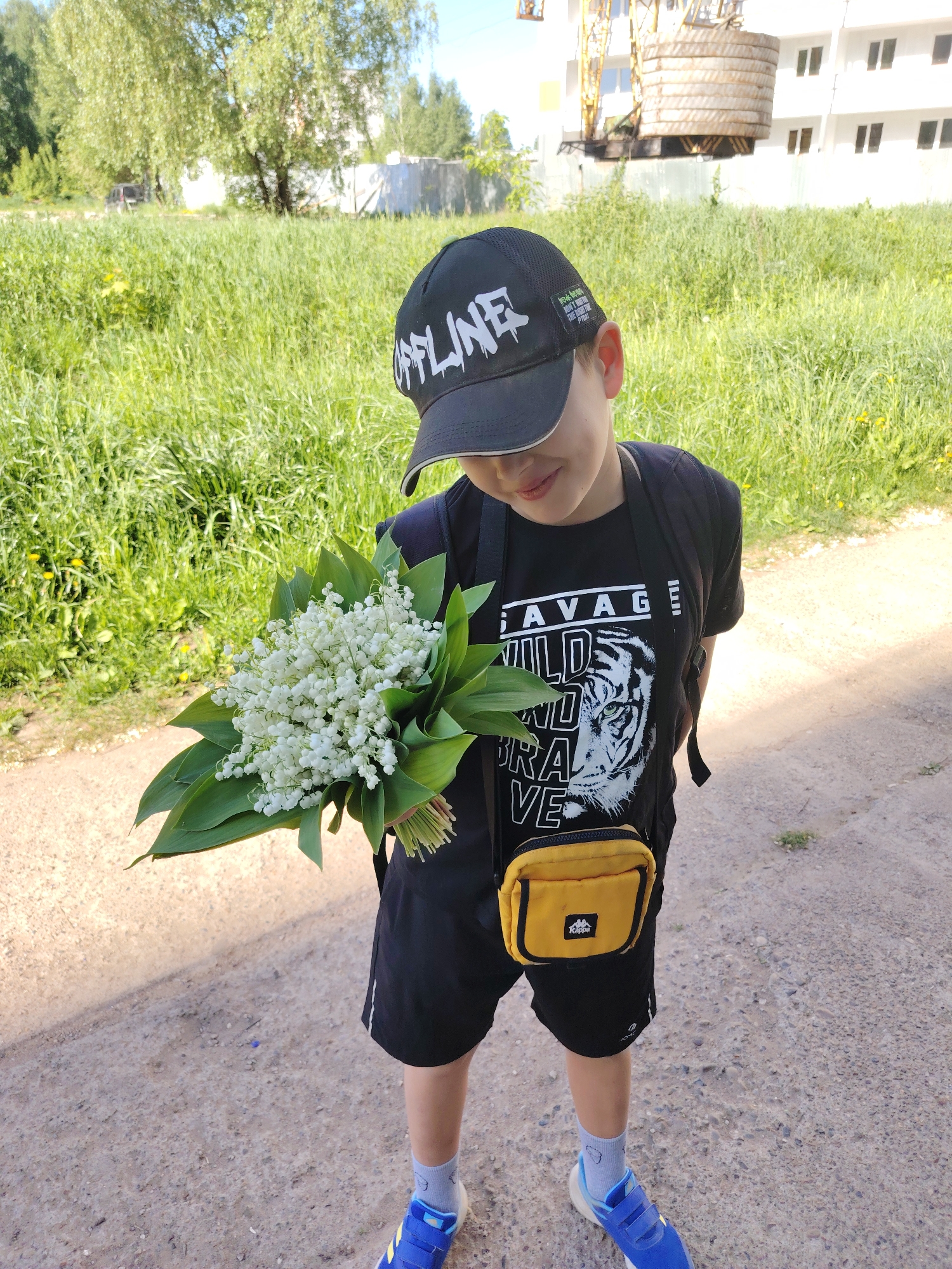 A young boy stands on a gravel path, holding a bouquet of lilies of the valley. He is wearing a black baseball cap with "OFFLINE" written on it, a black t-shirt with a tiger graphic and the words "SAVAGE WILD BRAVE", black shorts, blue sneakers, light blue socks, and a yellow crossbody bag. He also has a black backpack. The boy is looking down at the flowers with a slight smile.
The background includes a field of green vegetation, a metal fence, and a multi-story building under construction. A yellow crane is visible near the building.
The location appears to be in an urban area, possibly near a construction site. Based on visual clues, it could be Chaikovsky, Russia.