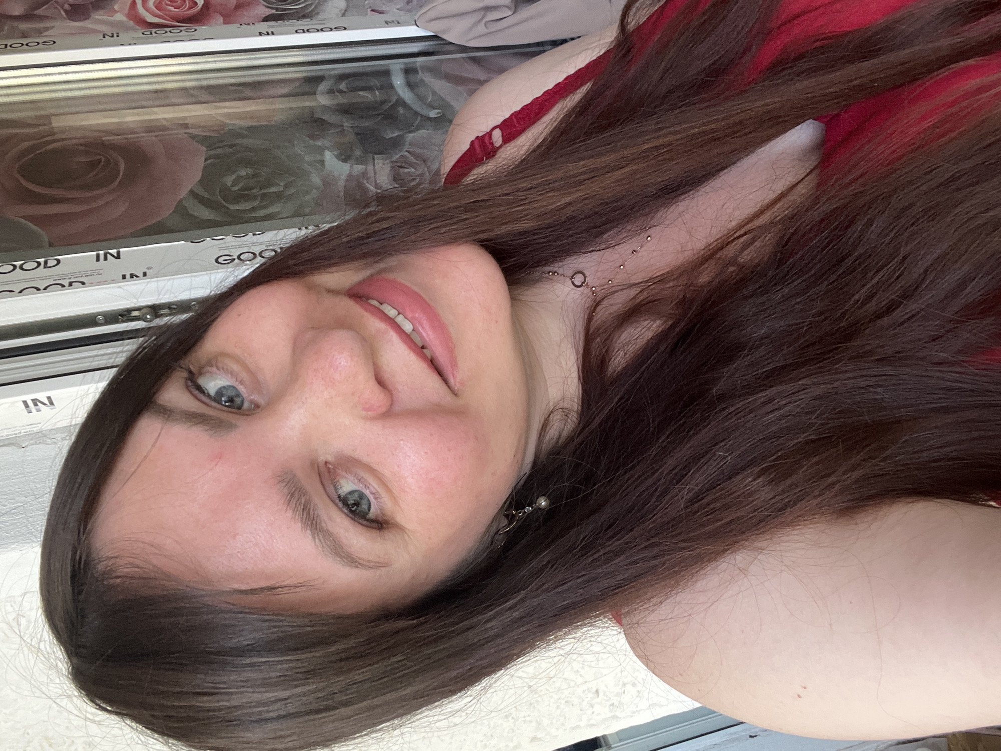 This media file is a selfie of a woman with long, dark brown hair and light blue eyes. She is wearing a red top and a necklace. Her face is partially obscured by her hair, and she is looking towards the camera with a slight smile. The background appears to be a window with a floral pattern and the text "GOOD IN" is visible on the window frame. The lighting suggests it is daytime. The image is rotated, with the woman's head at the bottom. The woman's expression appears relaxed and cheerful.
