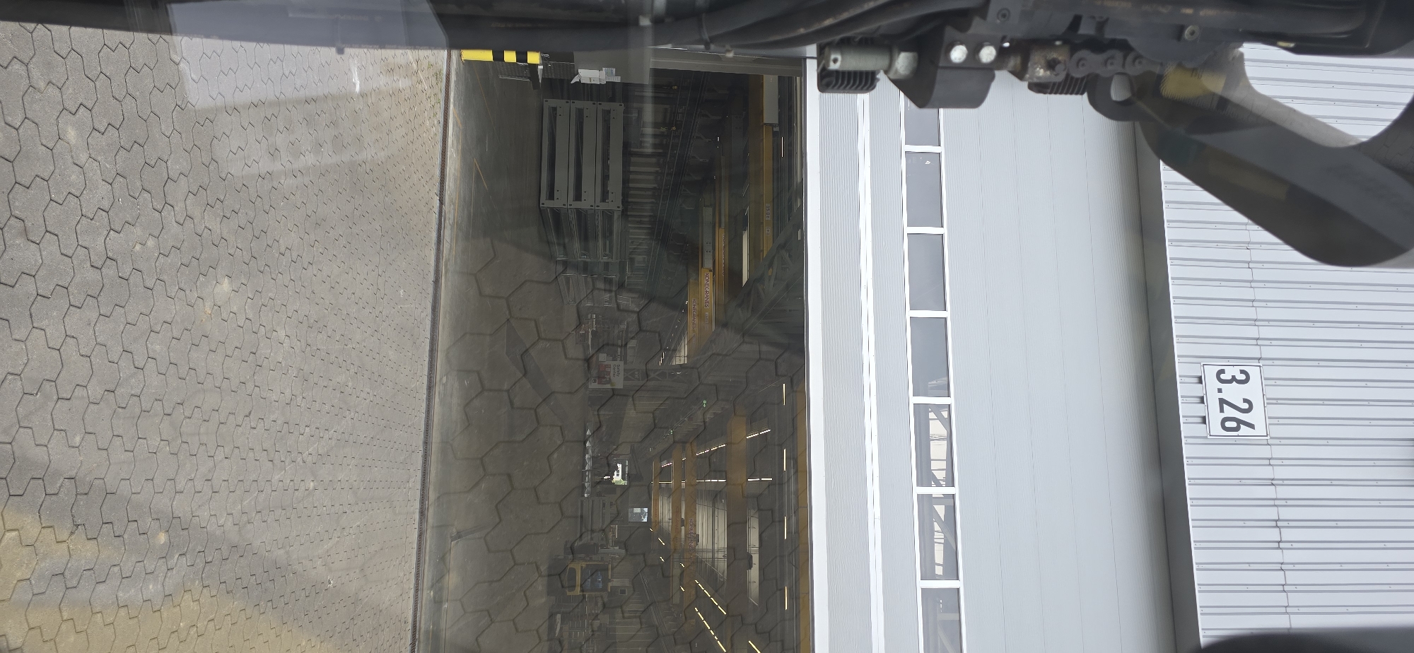 This is an outdoor, daytime image taken from a vehicle, likely a forklift or similar machinery, looking towards a large industrial building with a metal exterior. The foreground on the left is a gray, interlocking paver surface. Through a large window or opening on the left side of the building, an industrial interior is visible, filled with metal racks, yellow beams, and what appears to be some sort of manufacturing or storage equipment. The floor of the interior also looks like it's made of interlocking pavers.
On the right side of the image, the building has a corrugated metal facade. There are several tall, narrow windows with white frames. A white sign with the black text "3.26" is attached to the corrugated metal on the right side of the building. The overall scene suggests a logistics or manufacturing facility. There are no people visible. The lighting indicates it is daytime, and the weather appears to be fair. The location is identified as Haren (Ems), Germany.