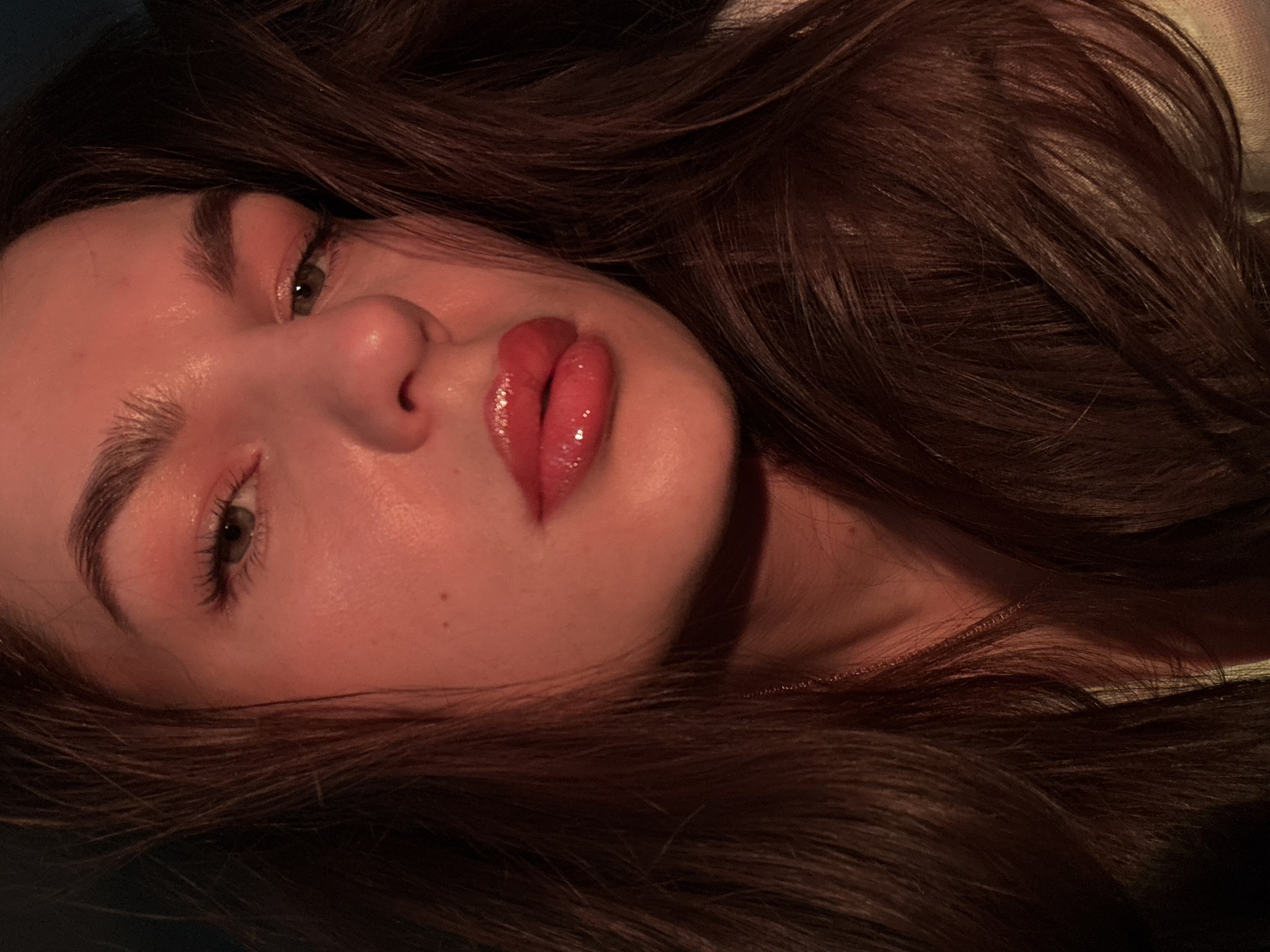 A close-up, slightly tilted shot of a young woman's face. She has fair skin, dark brown hair, full lips with a glossy red lipstick, and light-colored eyes with visible eyelashes. Her eyebrows are well-defined and slightly arched. The lighting is warm and muted, casting subtle shadows on her face. A thin gold chain is visible around her neck, and strands of her hair frame her face. The background is dark and indistinct. There is no indication that this image was taken in the city of Chaikovsky, Russia.