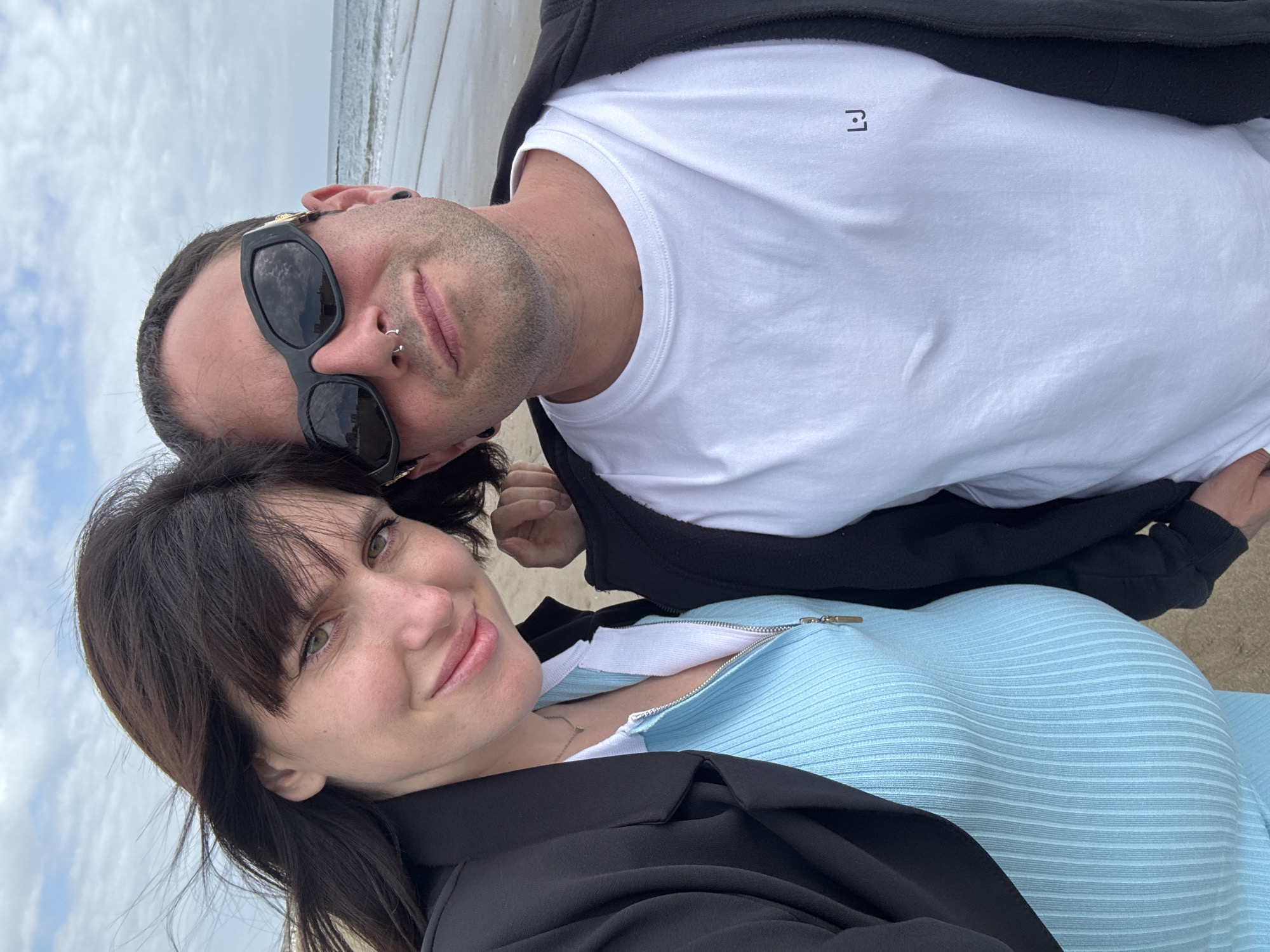 A selfie-style photograph, taken at an angle, captures a man and a woman on a beach in Nettuno, Italy. The background features the ocean with gentle waves and a cloudy sky, suggesting an overcast daytime.
The man, positioned towards the top-left of the image, wears black sunglasses that reflect the sky, a white t-shirt with a small, dark, stylized geometric logo on the left chest, and a dark jacket. He has dark hair, stubble, and visible facial piercings on his septum and nostril. His expression is neutral. The logo on his shirt appears to be an abstract design, possibly resembling an 'L' and a 'J' combined.
The woman, located towards the bottom-right, has dark hair with bangs and a subtle smile. She is wearing a light blue ribbed top with a silver zipper down the front, layered under a dark jacket. A delicate necklace is visible around her neck.
The overall scene conveys a casual and relaxed moment shared by the two individuals on the Italian coastline.