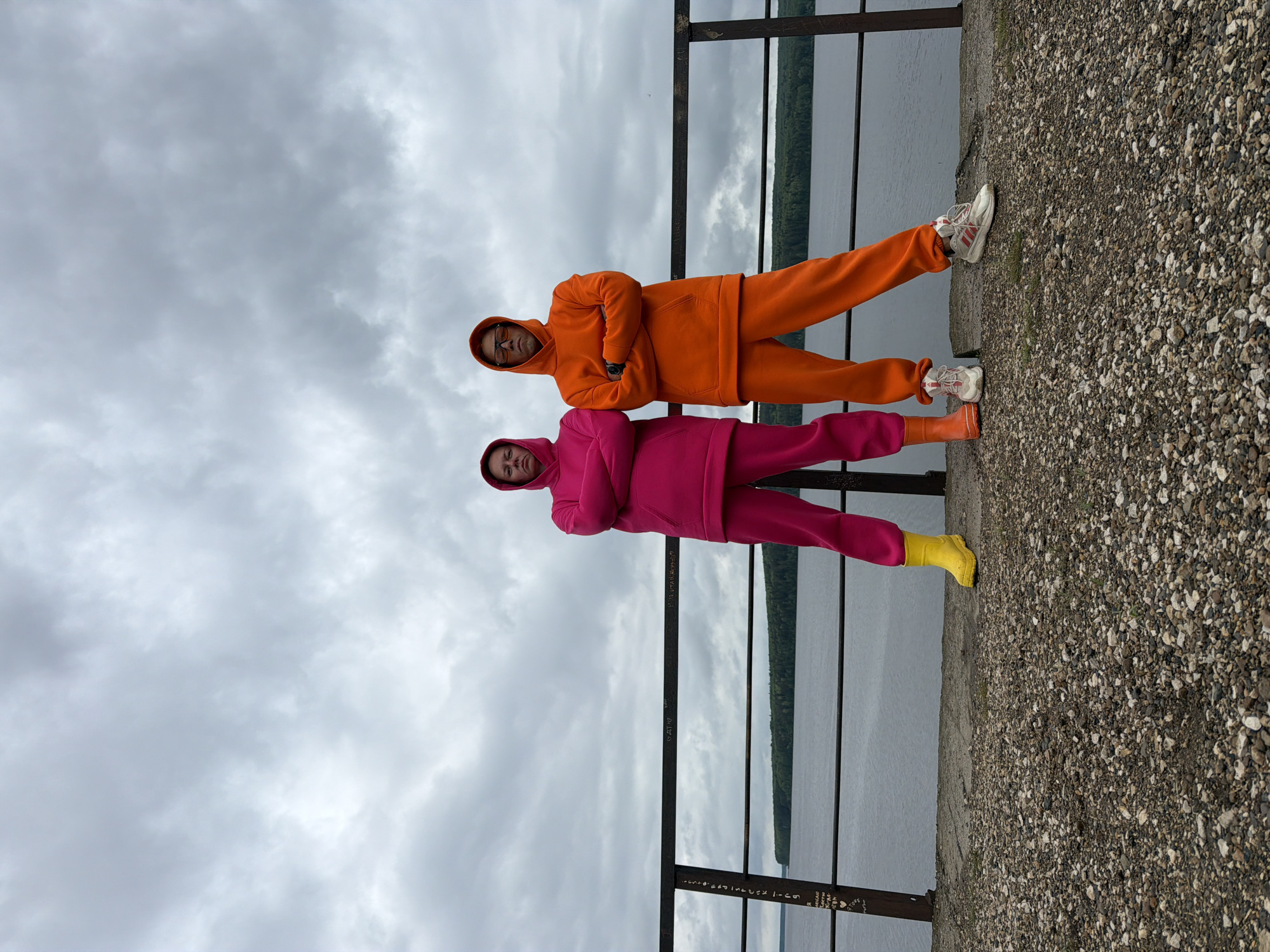Two individuals stand on a concrete structure overlooking a body of water, likely the Votkinsk Reservoir, within Chaikovsky, Russia, based on the Russian graffiti on the metal railing. The person on top wears an orange hoodie and pants, with white sneakers. The person below wears a pink hoodie and pants, with yellow boots. Both figures have their arms crossed and appear to be posed for the photograph. The background includes a cloudy sky, water, and a strip of land with trees. The surface they stand on is concrete and pebbles.