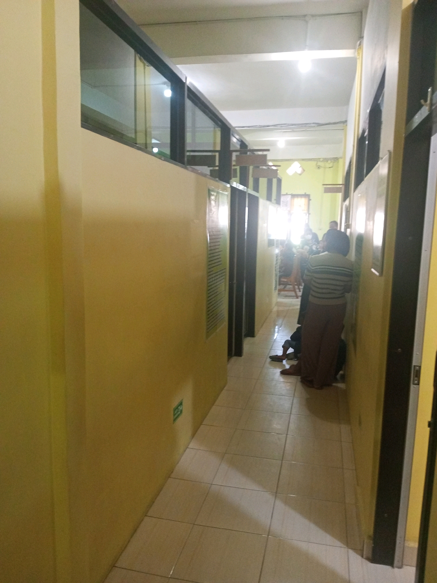 The image depicts a hallway with yellow walls and a tiled floor. The hallway is lit by fluorescent ceiling lights and natural light entering from an open doorway at the far end. There are several people visible in the hallway. In the foreground, on the right side, a person wearing a striped sweater and brown pants is standing. To the left and slightly behind this person, another individual is seated on the floor. Further down the hallway, several other individuals are visible, some seated and some standing, near a doorway. The walls have framed pictures or signs and a green exit sign is visible on the left wall. The overall scene suggests an indoor communal space, possibly an office or waiting area, within Kelurahan Kamalaputi, Indonesia.