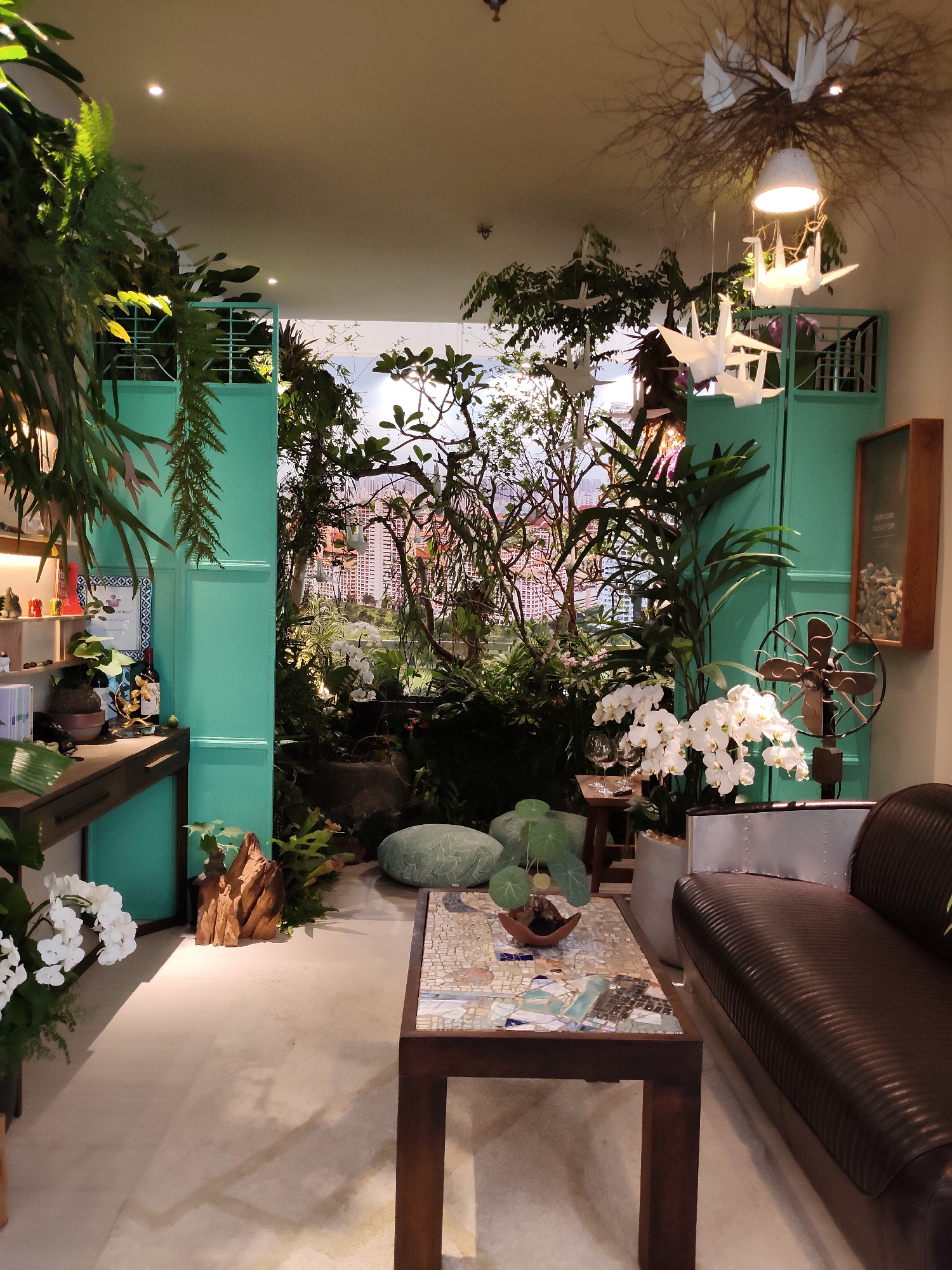 This is a photograph of an interior space decorated with plants and decorative objects. The scene appears to be a retail display or a well-styled living area. Lush green ferns and other plants hang from above and are arranged throughout the space, creating a jungle-like atmosphere. In the background, an open doorway reveals a panoramic view of a city skyline, suggesting a setting in a metropolitan area. The decor includes a dark leather sofa, a coffee table with a mosaic top, and several decorative items such as white orchids, a vintage fan, and origami cranes hanging from the ceiling. The overall aesthetic is a blend of natural elements and modern design. The scene does not depict any people. The backdrop of the city skyline suggests this location could be in a densely populated urban area.