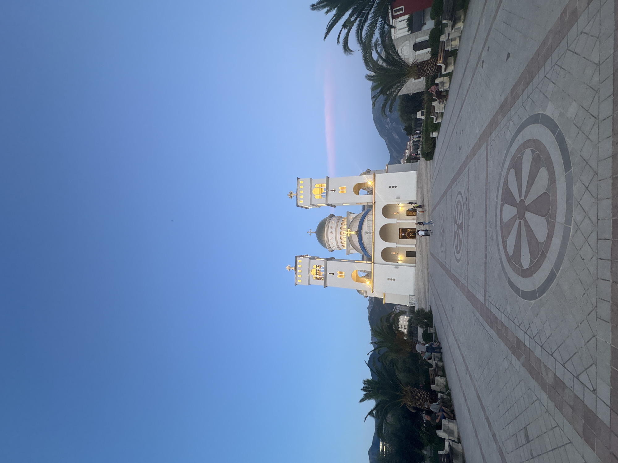 The image shows the Church of Saint John Vladimir in Bar, Montenegro. The church is a large, white building with two bell towers and a central dome. It is illuminated by lights, and the sky is a clear blue with a hint of pink from the sunset. In front of the church is a wide paved plaza with benches and palm trees. Several people are visible walking in the plaza and sitting on the benches. The scene is serene and peaceful, with the mountains in the background adding to the natural beauty of the location.