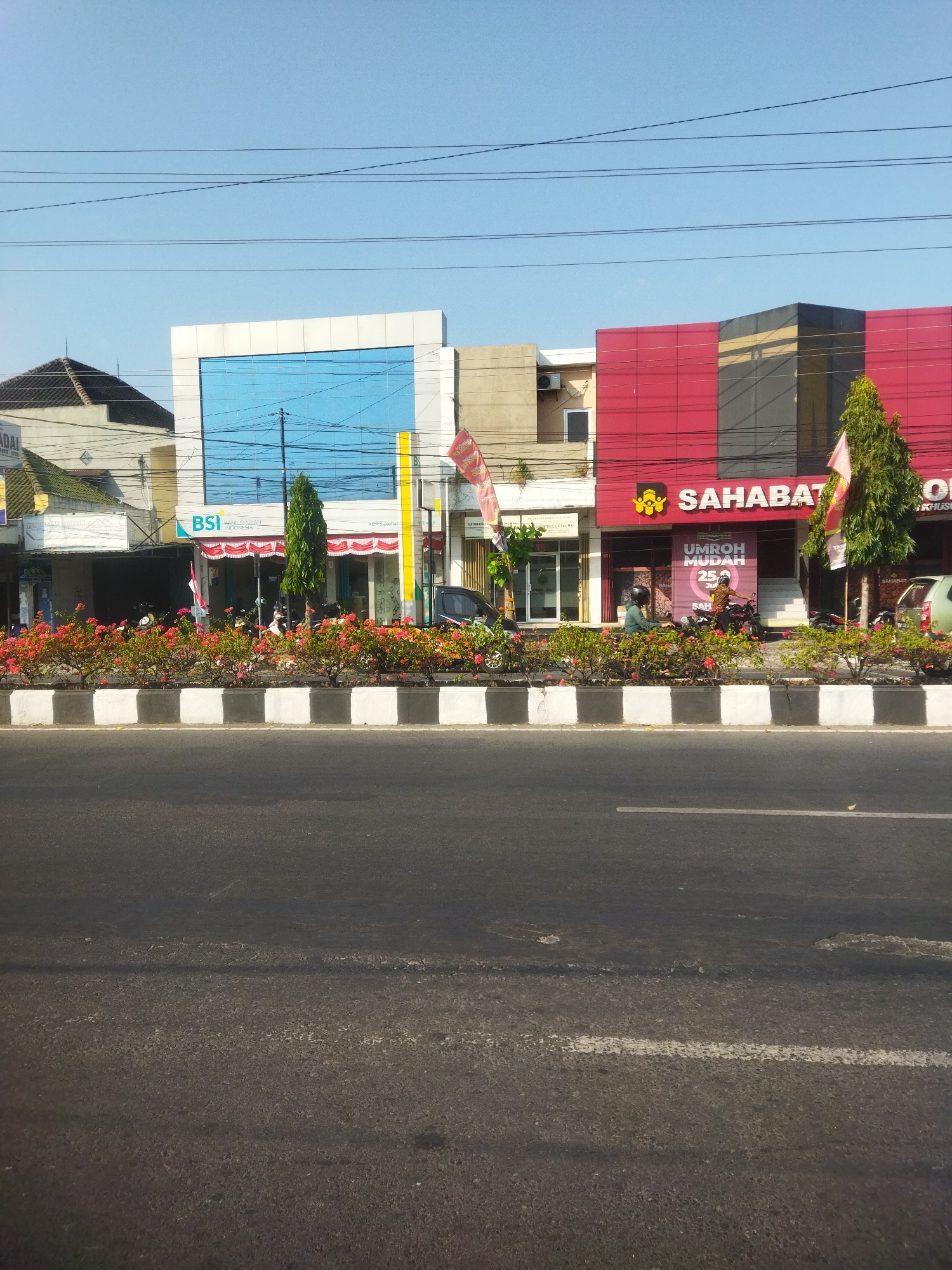 A street-level, eye-level, full-frame photograph captures a bright, sunny day in Sukoharjo, Indonesia. The scene features a row of commercial buildings lining a street. The buildings are varied in design and color. On the left, a building with a white facade and a large, light blue glass section is visible, with the "BSI BANK SYARIAH INDONESIA" logo prominently displayed. Next to it, a building with a red facade features the "SAHABAT" logo and advertising for "UMROH MUDAH". Adjacent to these, other buildings with simpler designs are present. A landscaped median strip filled with vibrant pink and red flowers separates the buildings from the asphalt road in the foreground. Several motorcycles are parked in front of the buildings, and a few people are visible, some on motorcycles. Electrical wires crisscross the sky above the buildings. The sky is clear and blue.