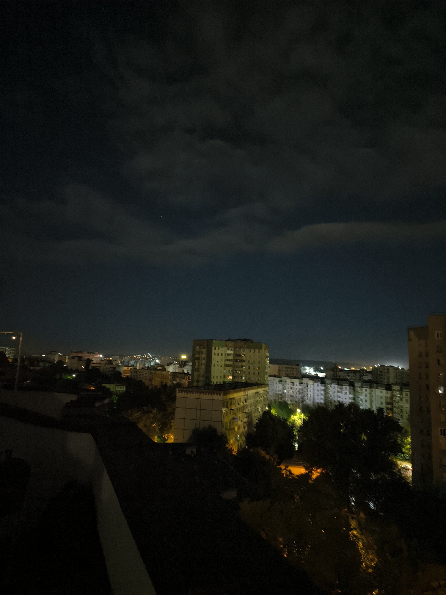 This image depicts a nighttime aerial view of a residential area in Chișinău, Moldova. The foreground features a dark rooftop or balcony railing. Beyond this, a densely populated urban landscape stretches into the distance, characterized by numerous apartment buildings of varying heights and architectural styles, many illuminated from within or by streetlights. Trees are interspersed throughout the cityscape, their foliage silhouetted against the artificial lights. The sky above is dark, with scattered clouds that are subtly illuminated by city lights, and a few faint stars are visible. The overall impression is one of a typical urban night scene in Chișinău.