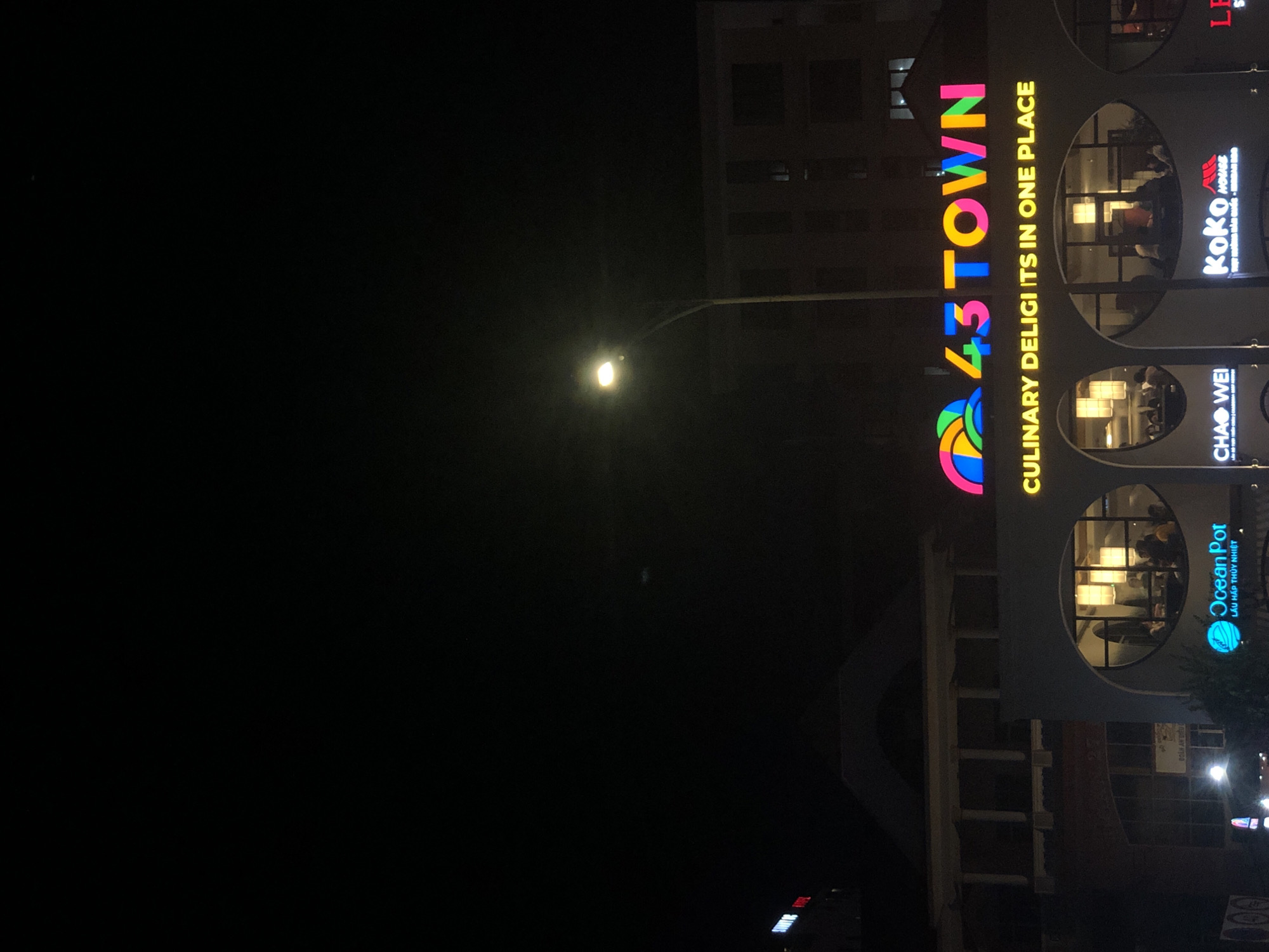This image captures a nighttime scene of a street in Da Nang, Vietnam, featuring the brightly lit sign for "43 TOWN Culinary Delights in One Place." The sign is characterized by its vibrant, multi-colored lettering, with "43 TOWN" displayed prominently in neon pink, yellow, blue, and green, and the tagline "CULINARY DELIGTS IN ONE PLACE" in yellow. Below the main sign, several businesses are visible, including "Ocean Pot" with its blue, globe-like logo and Vietnamese text, "CHAO WEI," and "KOKO HOUSE." The buildings in the background are illuminated, revealing architectural details such as arched windows and balconies. A streetlamp with a glowing bulb illuminates the foreground, casting a warm light on the scene. People can be seen inside the establishments, dining in dimly lit interiors. The overall atmosphere is that of a bustling commercial area at night.