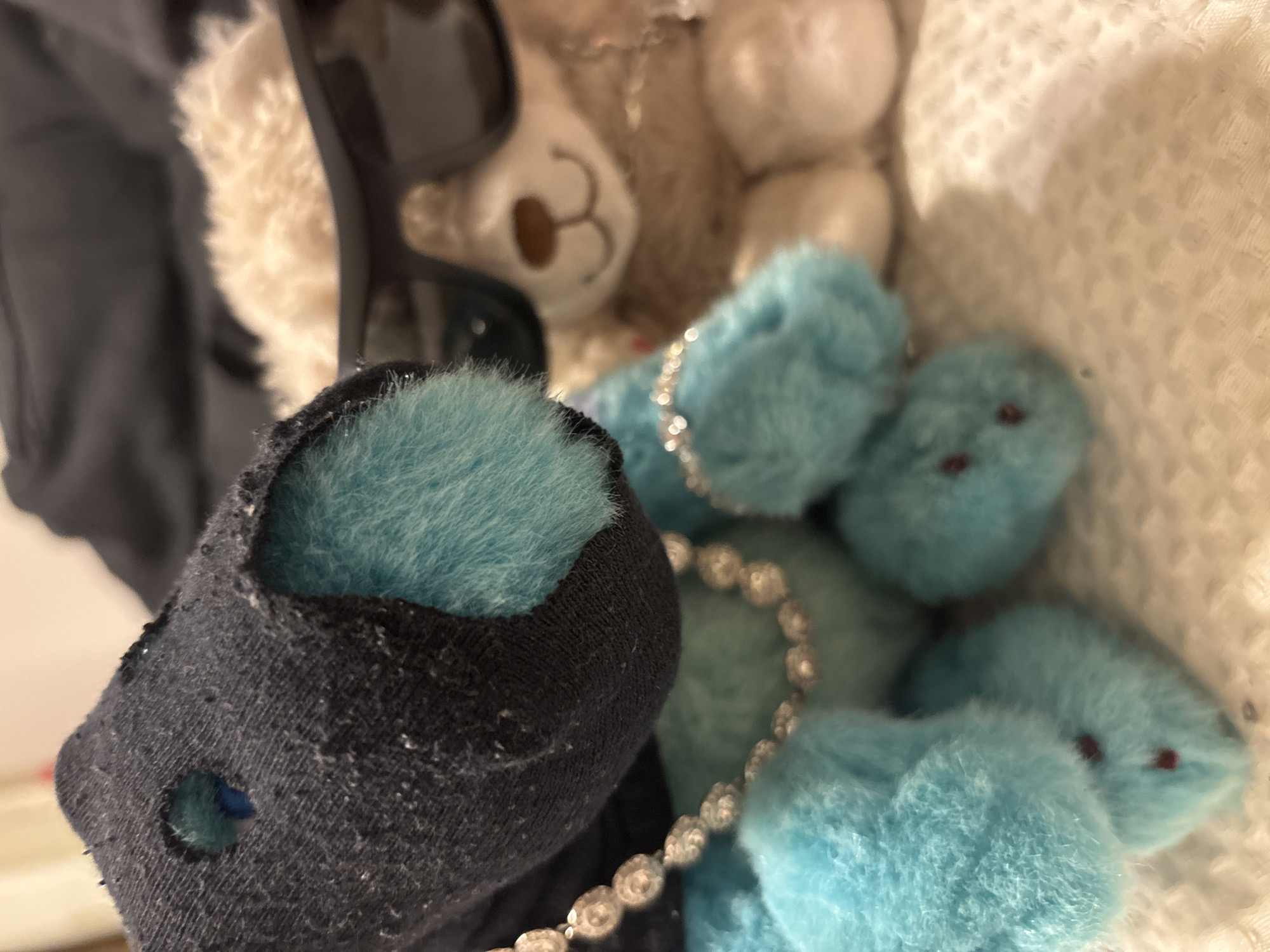 The image contains a close-up view of various objects. Dominating the foreground is a black sock with a turquoise, furry material visible within its opening. Sunglasses are positioned behind the sock, along with a white teddy bear. Further back, there are multiple fuzzy, blue pom-poms with small red eyes and a beaded chain. The setting is not explicitly identifiable as Buenos Aires, Argentina, and there are no people, actions, or interactions apparent in the frame. The environment is undefined beyond the objects themselves.