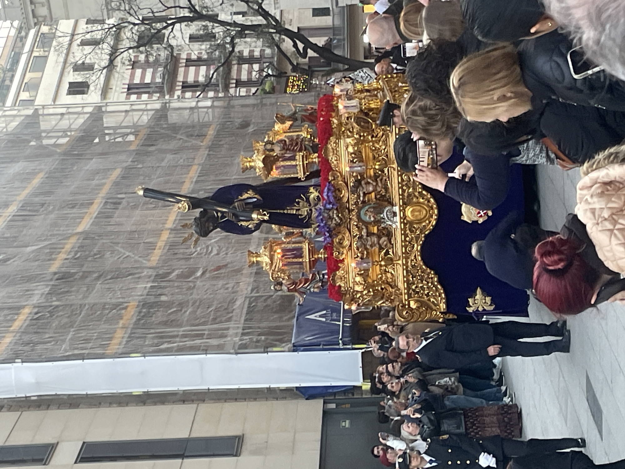 The image depicts a religious procession taking place on a city street in Barcelona, Spain. A large, ornate golden platform bearing a statue of Jesus carrying the cross is surrounded by a crowd of onlookers. Many in the crowd are capturing photos or videos with their phones. Building undergoing construction can be seen behind the procession.
