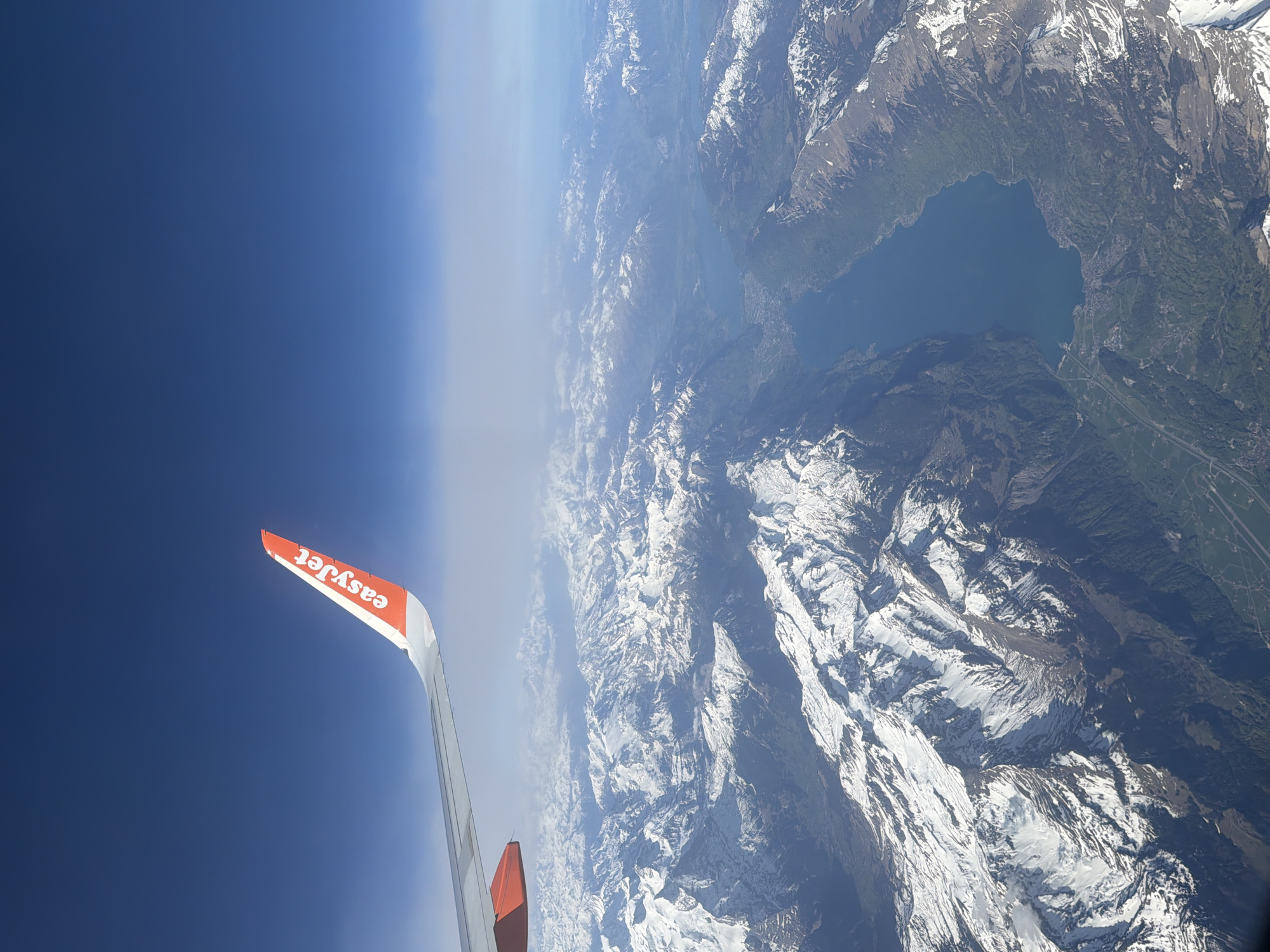 An aerial photograph captures a breathtaking view from an airplane window, looking out over a vast mountainous landscape. In the lower-left foreground, the prominent orange and white wingtip of an EasyJet aircraft is clearly visible, extending diagonally upwards and to the right. The airline's name, "easyJet," is distinctly written in white text on the orange portion of the wingtip.
Below the aircraft, an extensive range of rugged, snow-capped mountains dominates the scene. Many of the peaks and higher slopes are covered in bright white snow, which contrasts sharply with the darker rock faces and patches of green forest and vegetation in the valleys and lower elevations. A large, deep blue lake is nestled amidst the mountains in the mid-ground, its calm surface reflecting the clear sky. Along the shores of the lake and within the adjacent valleys, faint signs of human presence, such as roads and buildings, can be discerned.
The sky, occupying the upper left of the image, transitions from a deep, clear blue directly above to a lighter, hazy blue closer to the horizon on the far right. This atmospheric haze adds depth to the distant mountain ranges. The scene is illuminated by bright daylight, suggesting a clear late morning or early afternoon. The overall impression is one of immense scale and natural beauty, consistent with a flight over the European Alps, given the flight context of Milan, Italy.