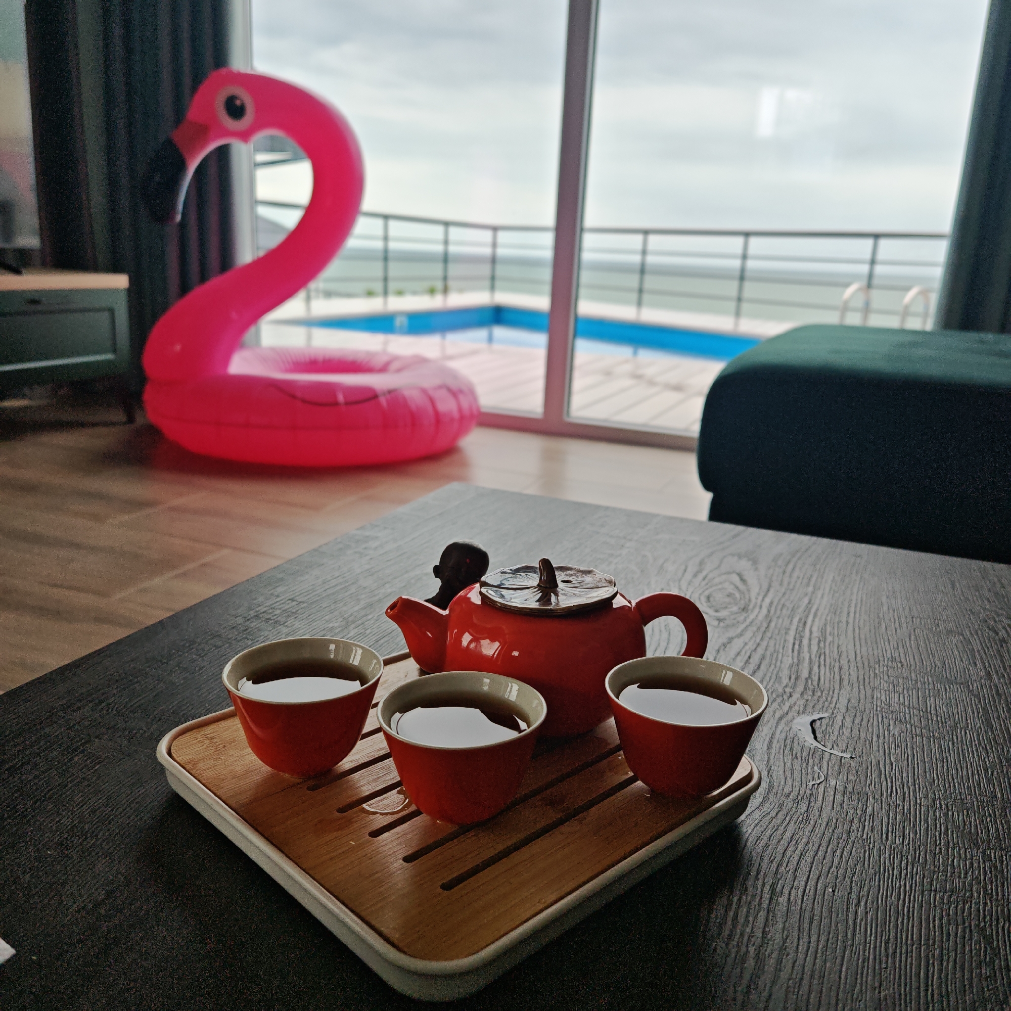 A still life scene within a room, likely a vacation rental or hotel room in Мысовское сельское поселение, Ukraine, features a wooden coffee table holding a red teapot and three matching teacups on a light-colored wooden tray. The cups and teapot contain a dark liquid, presumably tea. Behind the table, an inflatable pink flamingo is visible near sliding glass doors, revealing an outdoor pool area with a view of the sea under a cloudy sky. A green sofa or ottoman is also partially visible to the right of the glass doors. The room has tiled flooring. The scene presents a serene and relaxing atmosphere, indicative of a vacation setting.
