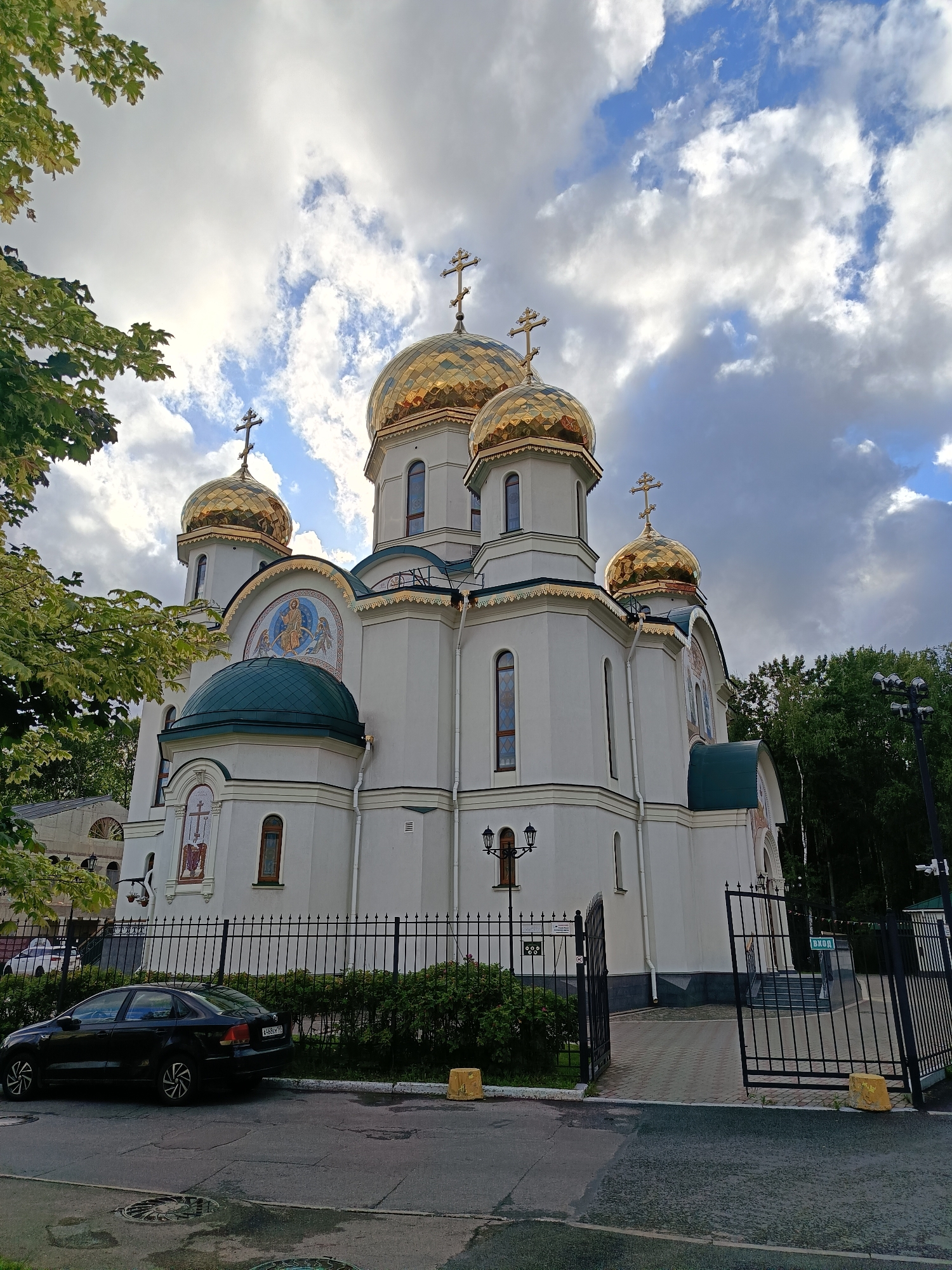 The image shows the Church of the Icon of the Mother of God "Joy of All Who Sorrow" in Saint Petersburg, Russia, on a cloudy day. The church is a white building with multiple golden domes, each topped with a cross. One large green dome is visible. The church is surrounded by a black metal fence, and lush green trees and bushes are present. In the foreground, a black car is parked on the side of a wet asphalt road. The sky is filled with dramatic clouds, with patches of blue sky visible. A street lamp and a security camera are also visible in the image.