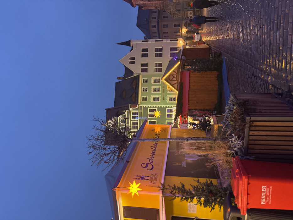 A European town square at dusk features cobblestone streets and historic buildings. A Christmas market is set up with stalls decorated with lights and signage for "Schallbenfest." A red NESTLER trash bin is visible in the foreground, along with a small fir tree. A few people walk in the distance.