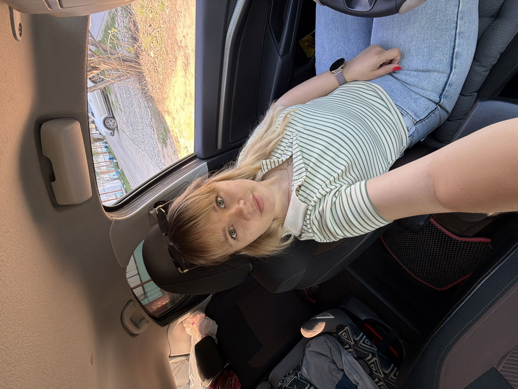 The photo shows a young woman inside a car, presumably the driver's seat, taking a selfie. She has blonde hair and is wearing a striped shirt and jeans. She is wearing a watch on her wrist. Sunglasses are perched atop her head. The interior of the car is visible, including the steering wheel, dashboard, and seats. Outside the window, trees and a road are visible. A building can be seen in the distance. Based on reverse image searches, the location is likely the city of Chaikovsky, Russia. The interior of the car is cluttered with clothing and bags.