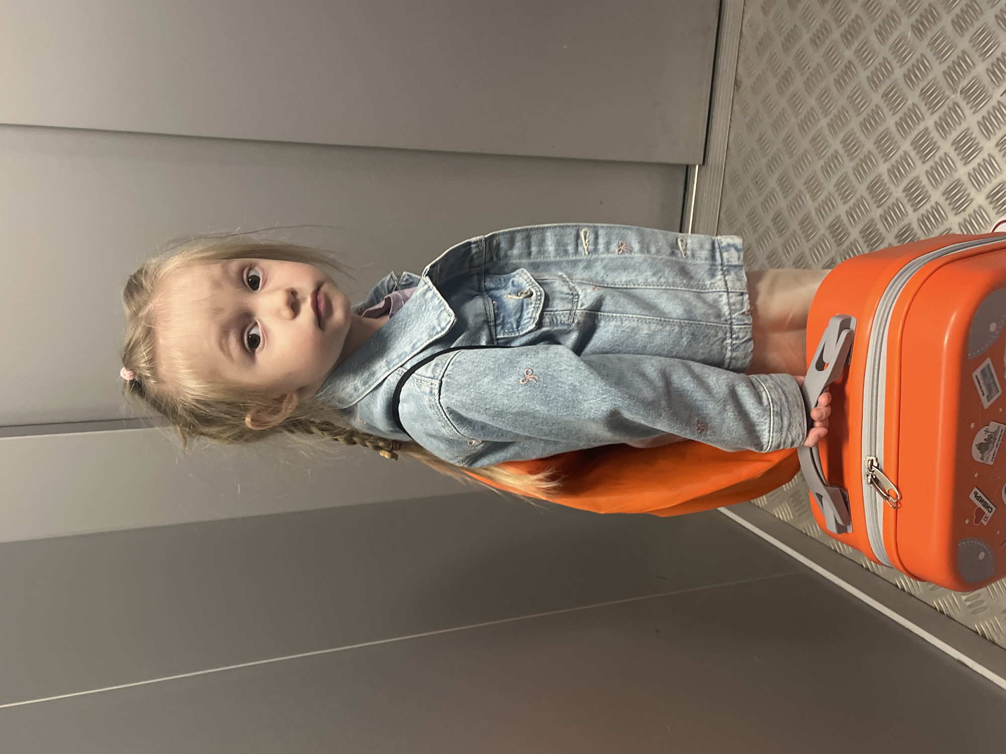 A young girl with blonde braided hair and a pink hair tie stands holding an orange suitcase. She is wearing a light blue denim jacket over a purple shirt and light-colored shorts. The girl is positioned in what appears to be an elevator, indicated by the metallic checkered floor and gray walls. Her expression is directed towards the camera. The background is a plain gray wall, likely part of the elevator interior. There are no specific visual cues to indicate the city of Chelyabinsk, Russia, in this image.