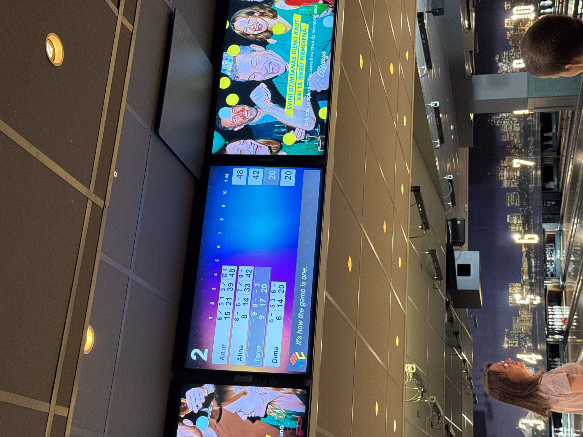 The image presents a forensic view of an O'Learys bowling alley in Riga, Latvia. The scene is illuminated by numerous recessed circular yellow lights embedded in a ceiling composed of acoustic panels. Various pieces of ceiling-mounted equipment, including speakers and sensors, are visible.
In the lower-left foreground, a woman with long brown hair and eyeglasses, wearing a light-colored short-sleeved top, stands facing right, observing the bowling lanes. In the lower-right foreground, the back of a person's head with short brown hair is partially visible, also oriented towards the lanes.
Above the bowling lanes, two prominent digital screens display information. The left screen shows bowling scores for four players: Artur (48 points), Alina (42 points), Tanja (20 points), and Dima (20 points), with individual frame scores displayed up to the fifth frame. A logo and the text "It's how the game is one." are visible at the bottom. The right screen features a promotional advertisement for O'Learys, showing four adults celebrating with drinks, against a backdrop of yellow polka dots. Latvian text on this screen translates to "CELEBRATE BIRTHDAY EVEN IF IT FALLS ON MONDAY" and identifies "O'Learys entertainment complex."
The background reveals multiple bowling lanes with pins set up. Behind the lanes, a dark-toned mural or projection depicts a cityscape, with large, illuminated numbers (e.g., '5', '6', '7') integrated as lane markers.